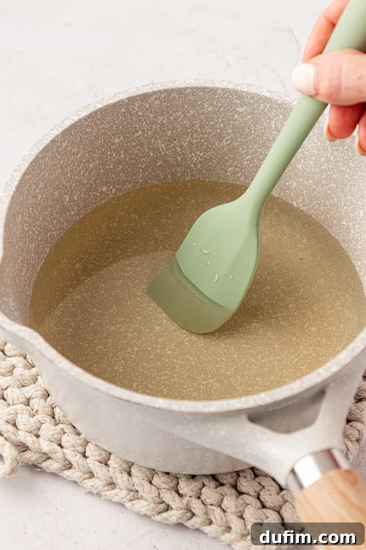 A hand gently stirs a clear liquid in a gray saucepan with a green silicone spatula, set on a beige woven mat. The mixture has transformed from cloudy to clear, indicating the sugar has dissolved.