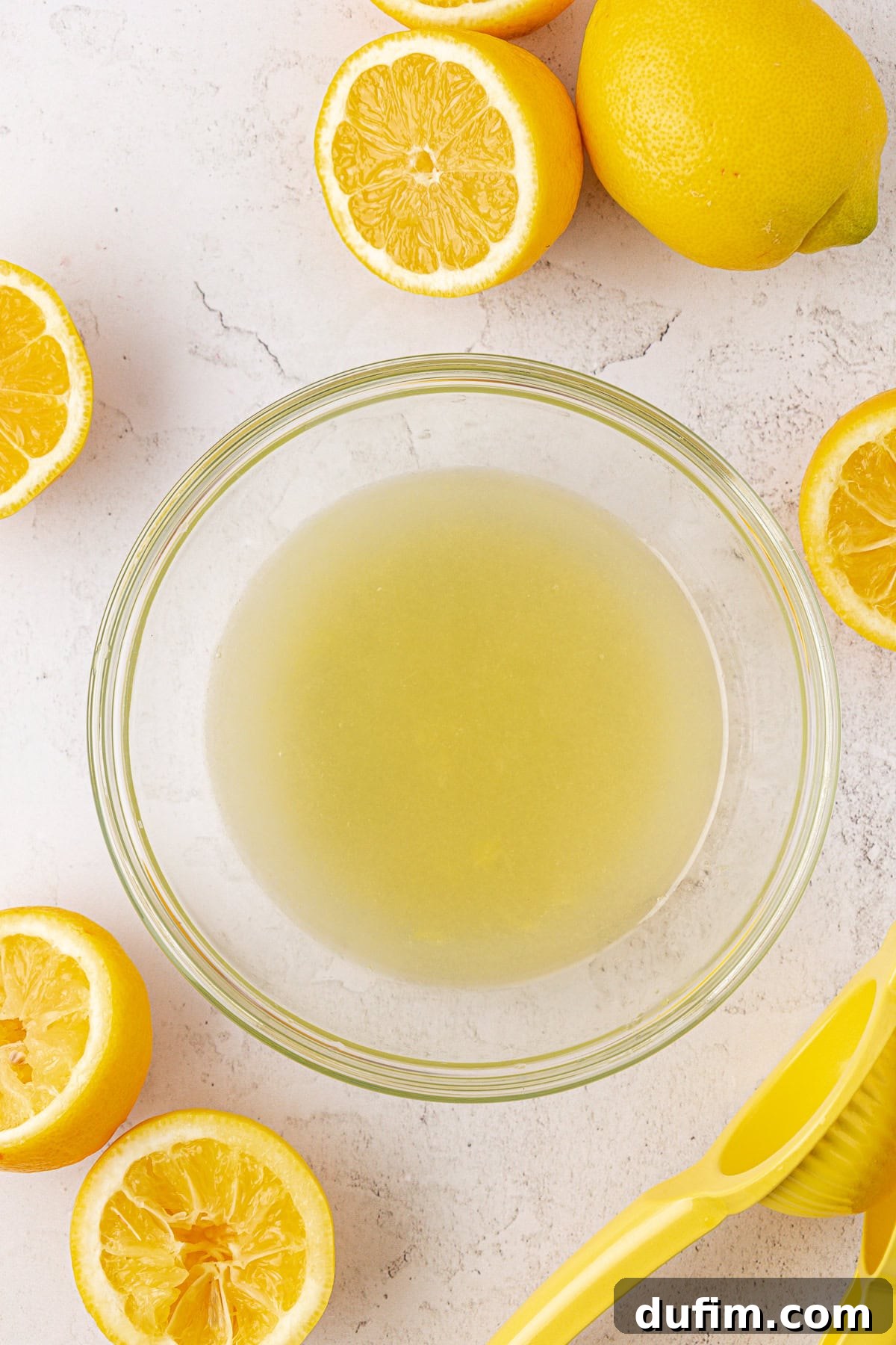 A clear glass bowl brimming with freshly squeezed lemon juice, surrounded by several whole and halved lemons on a light surface. A yellow citrus juicer waits nearby, ready for more juicing.