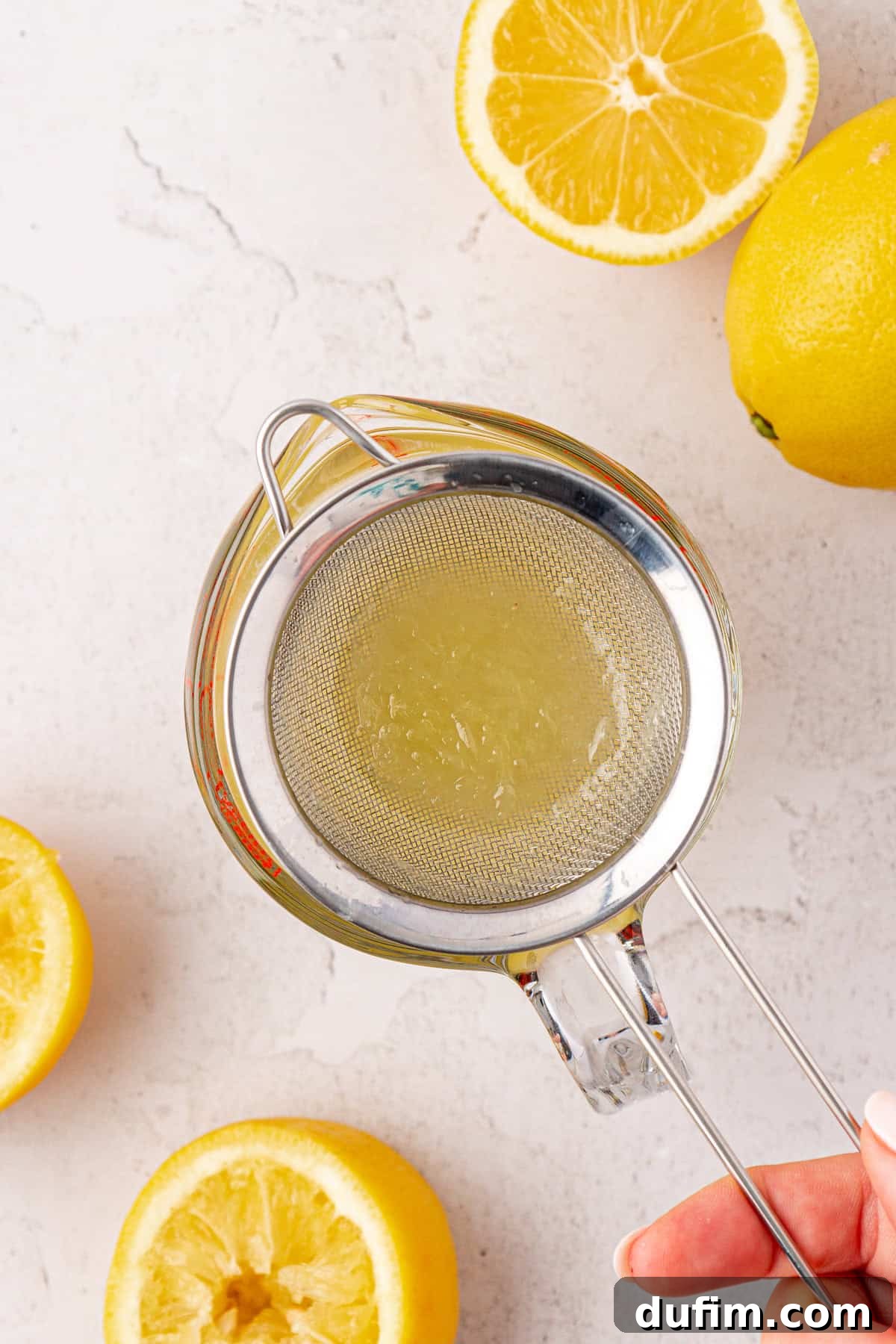 A hand holding a metal strainer over a glass jar, diligently filtering lemon juice to remove seeds and pulp. Halved and whole lemons are scattered across a white surface, indicating the juicing process is underway.