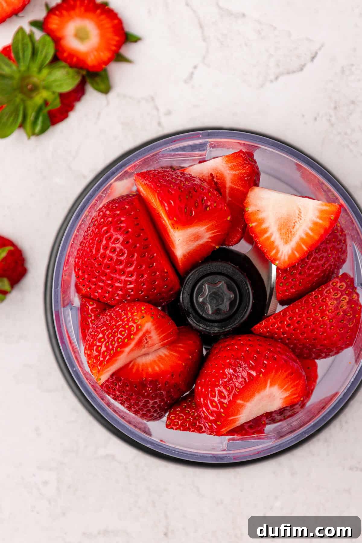 Fresh, vibrant strawberries, some whole and some halved, are carefully arranged inside a small food processor bowl on a light-colored surface. More whole strawberries and green leaves are nearby.
