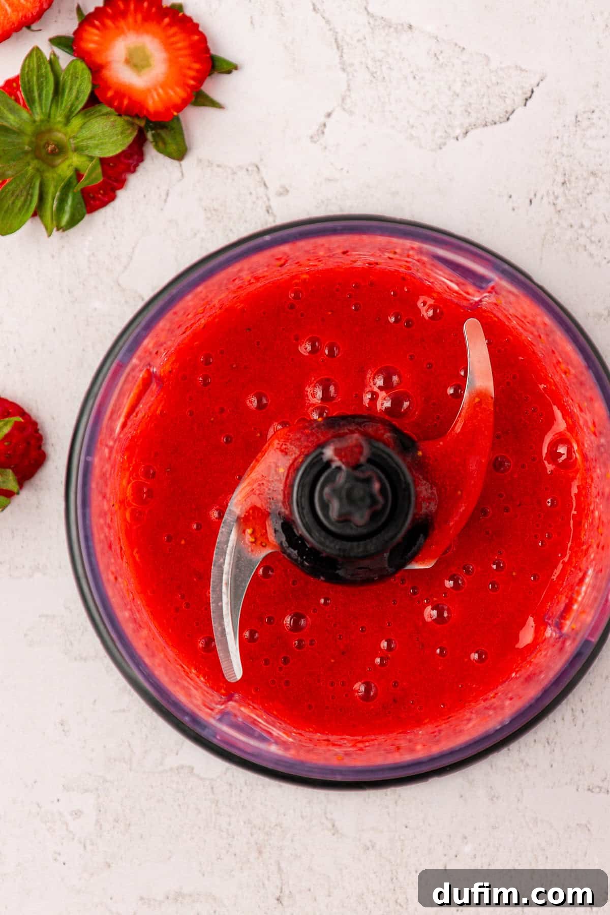 A food processor bowl filled with smooth, vibrant, bright red strawberry puree. Whole and sliced fresh strawberries are scattered on a light-colored surface beside the bowl.