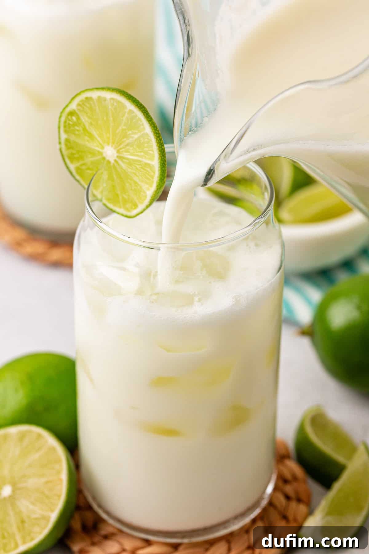 Crafting Creamy Brazilian Lemonade 2 A glass filled with ice is being poured with a creamy white drink from a pitcher. The glass is garnished with a slice of lime, and whole and cut limes are placed nearby on a coaster.
