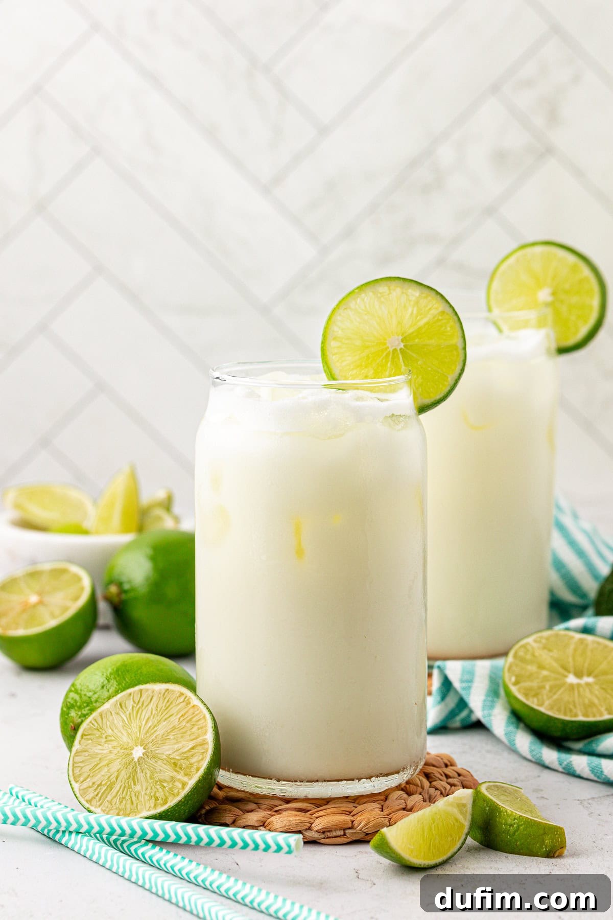 Crafting Creamy Brazilian Lemonade 11 Two glasses of a creamy, iced white drink garnished with lime slices, surrounded by fresh cut limes and blue striped straws on a light surface with a white tiled background.