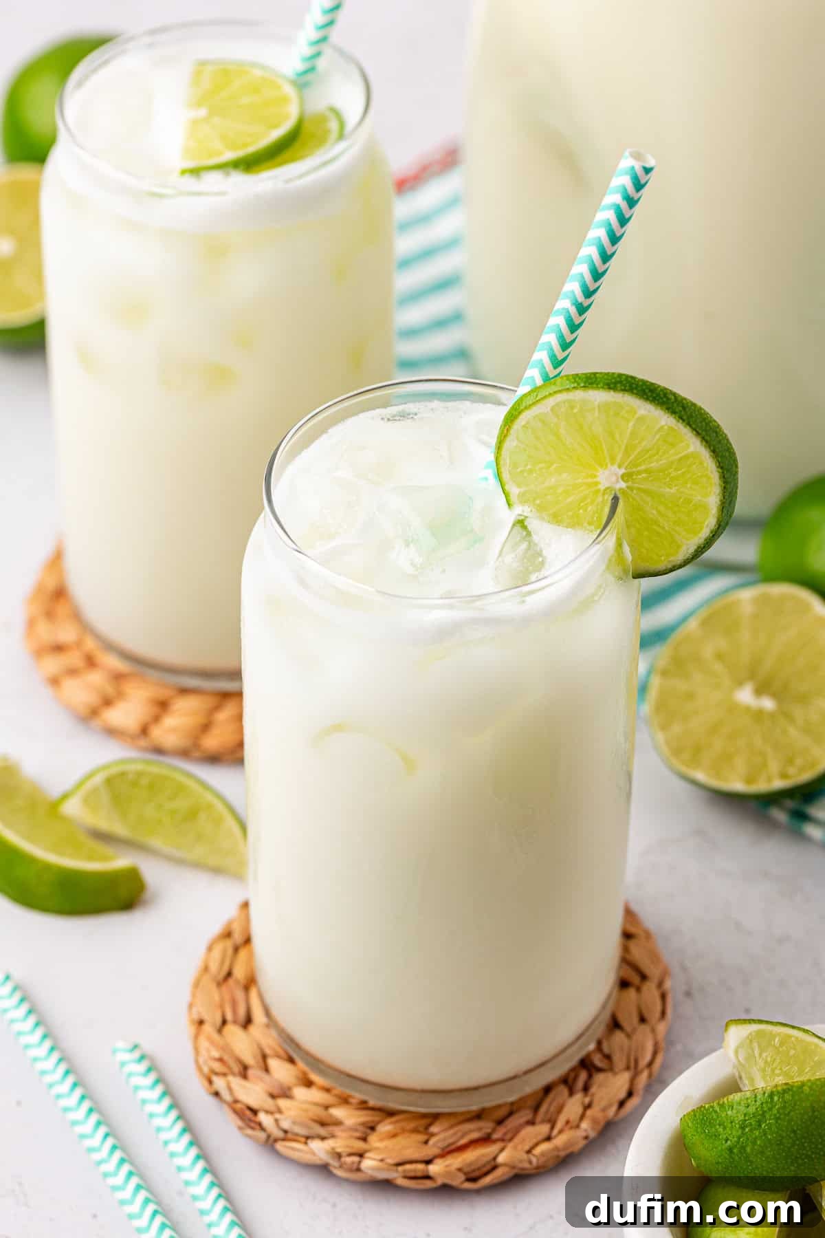Crafting Creamy Brazilian Lemonade 12 Two glasses of creamy, iced limeade with green-and-white striped straws, garnished with lime slices, sit on woven coasters. Fresh lime wedges are scattered around on a light surface.