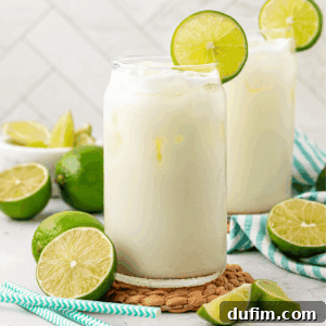Two glasses of a creamy white drink with ice, each garnished with a lime slice, sit on a woven mat. Fresh limes and blue-striped straws are nearby, with a white tiled background.
