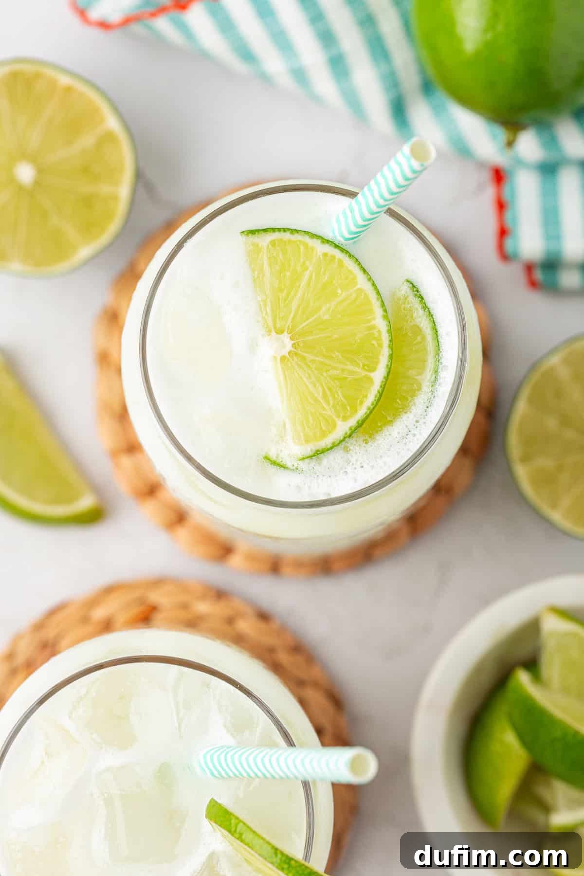 Crafting Creamy Brazilian Lemonade 4 Two glasses filled with ice and a light, frothy drink, each garnished with lime slices and a striped straw, surrounded by additional lime wedges on a white surface.