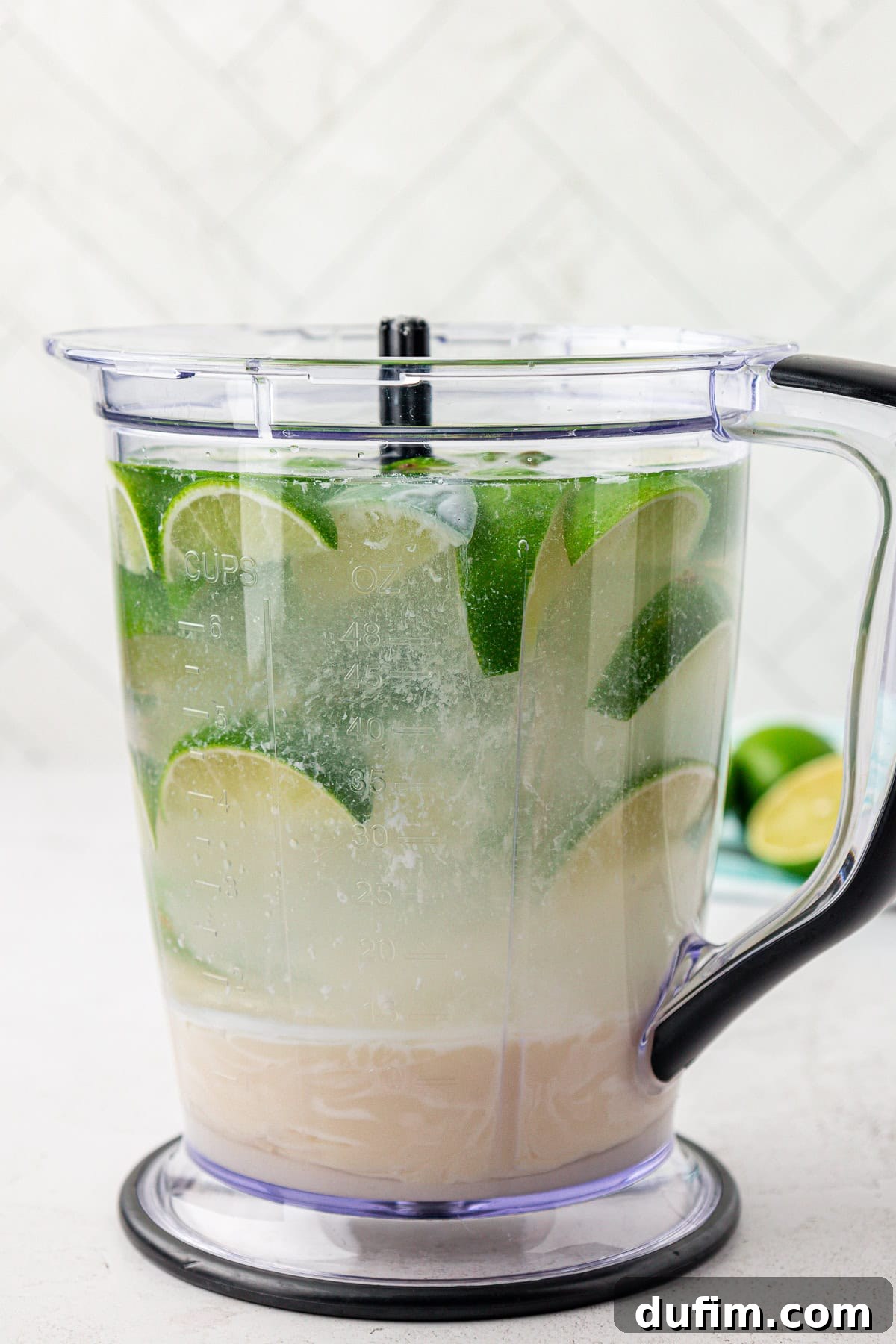 Crafting Creamy Brazilian Lemonade 5 A blender pitcher filled with water, lime wedges, and a cloudy liquid, sitting on a white surface with a white tiled background.