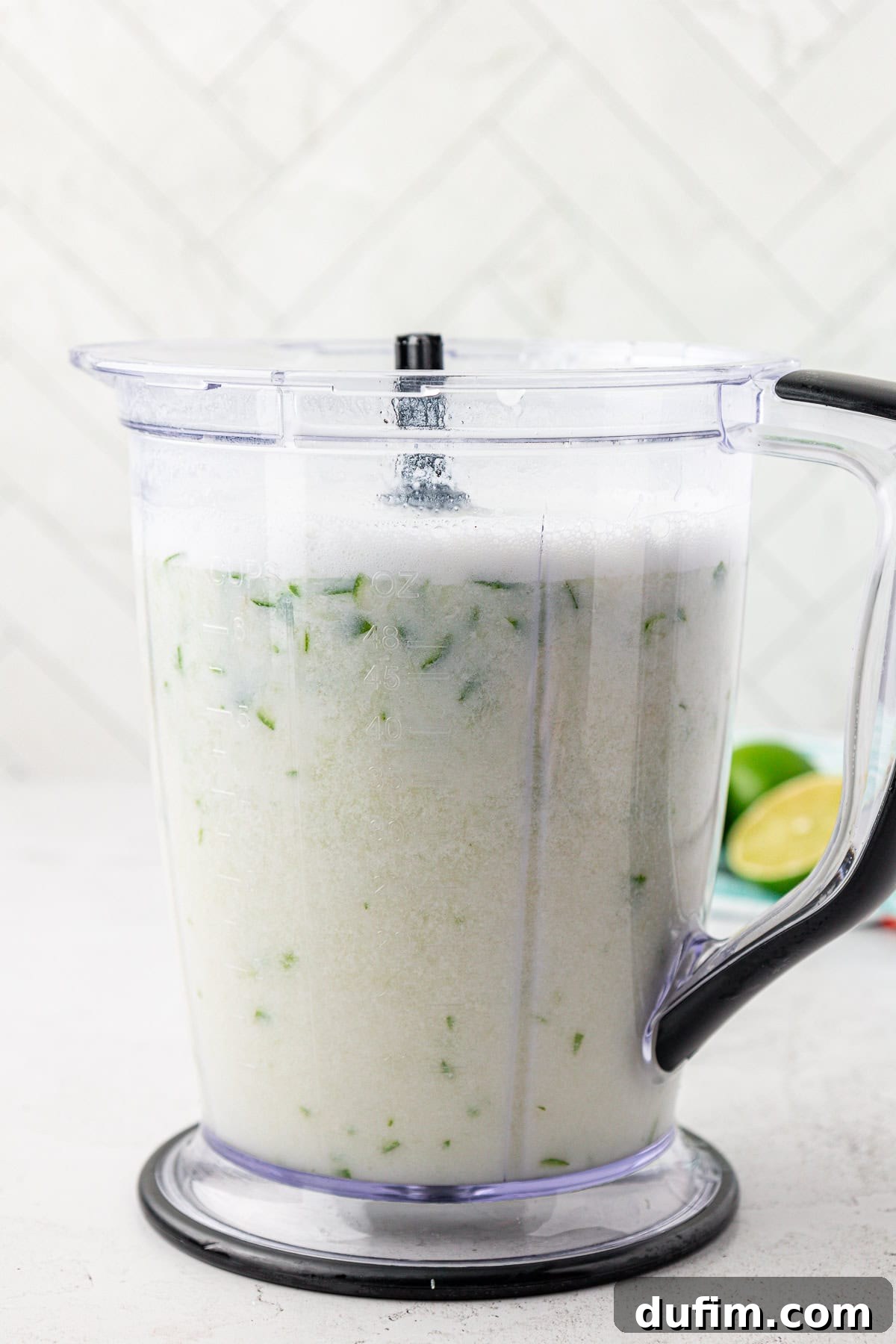 Crafting Creamy Brazilian Lemonade 6 A blender filled with a frothy, light-colored liquid containing small green pieces, sitting on a white surface. Lime halves are visible in the background.