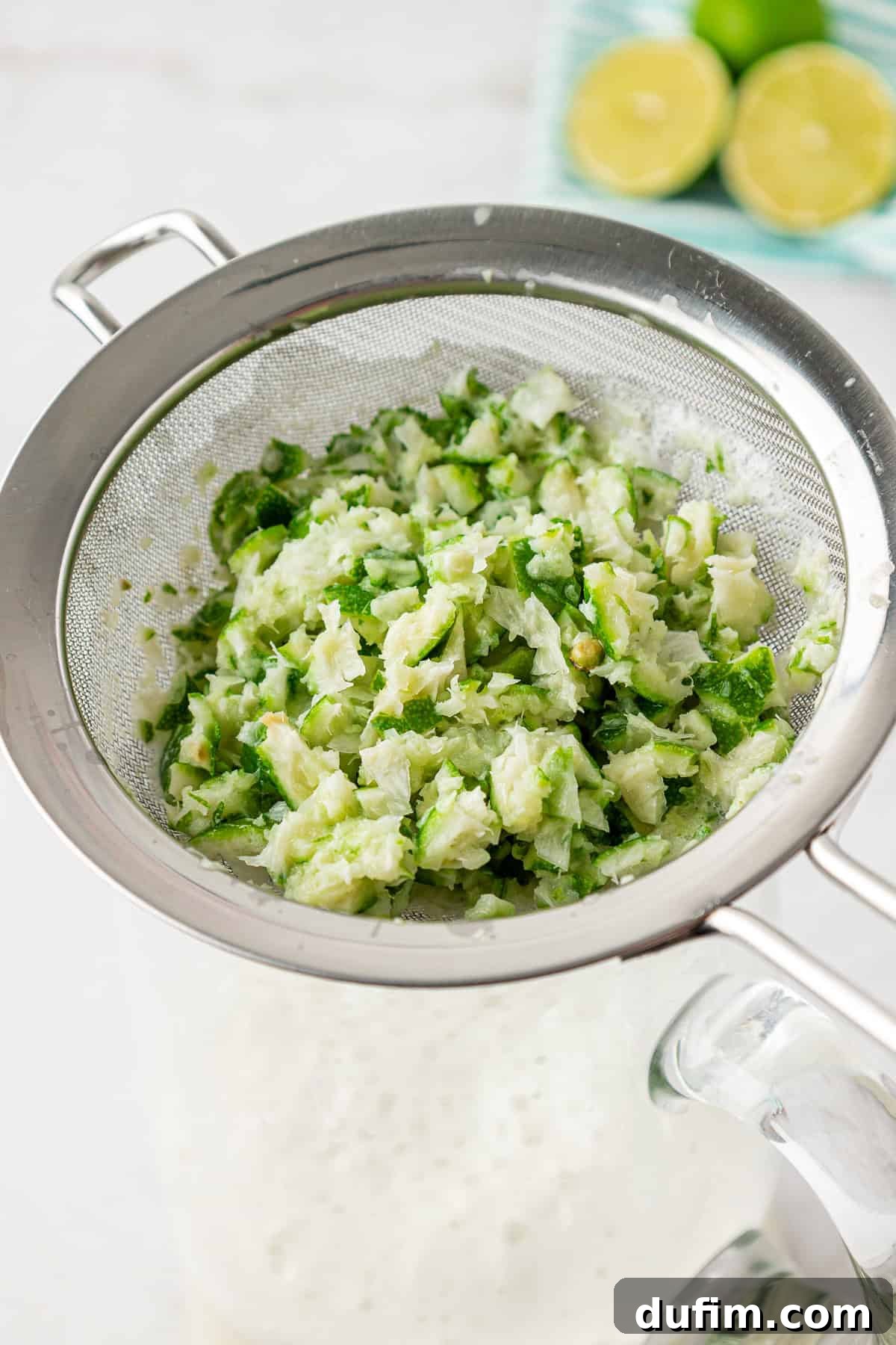 Crafting Creamy Brazilian Lemonade 8 A metal strainer filled with finely chopped green and white vegetables sits atop a glass pitcher. In the background, there are halved limes on a plate.