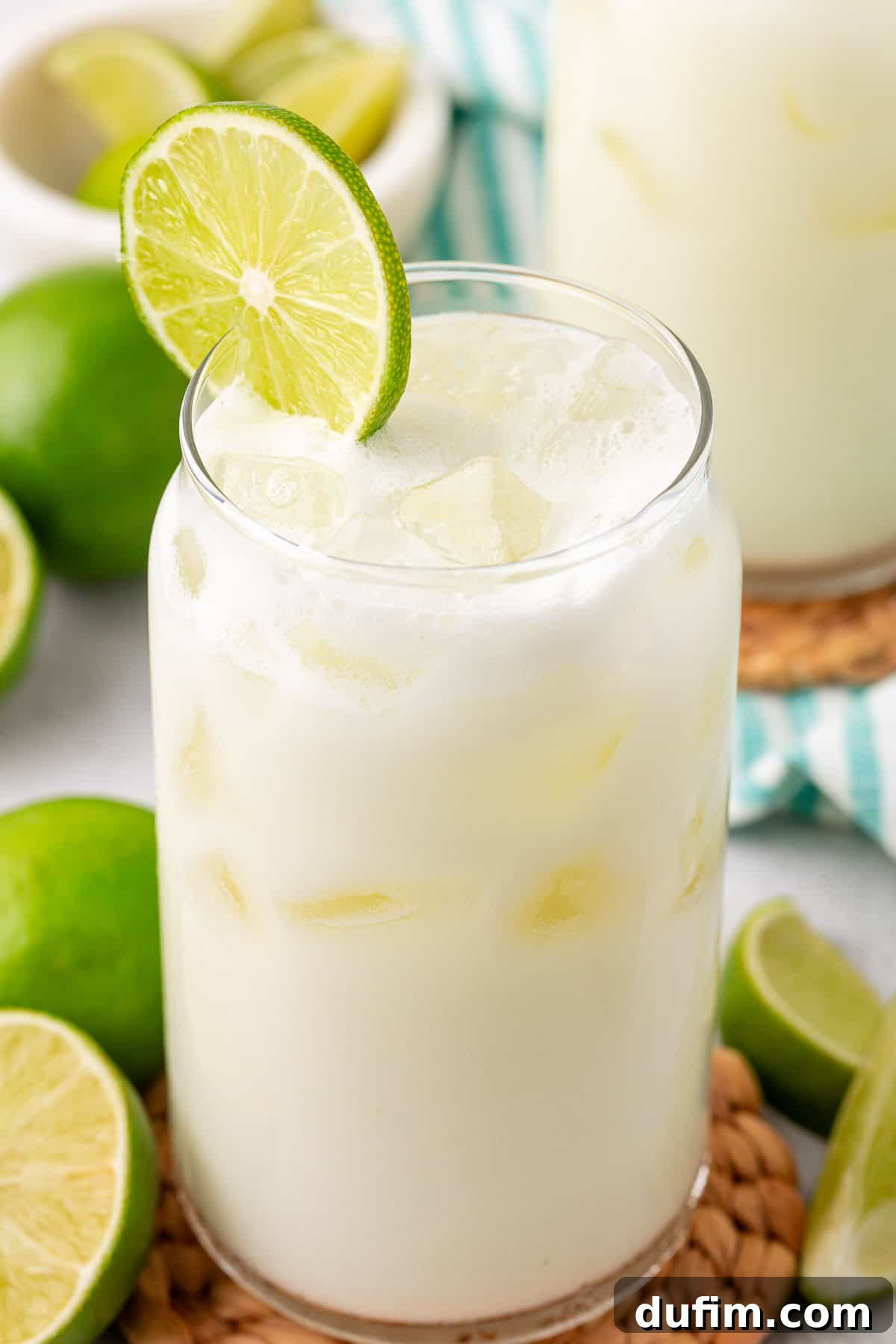 Crafting Creamy Brazilian Lemonade 9 A glass of iced, creamy white lime drink garnished with a slice of lime sits on a woven coaster. Fresh limes and lime wedges surround the glass, and a bowl of limes is in the background.