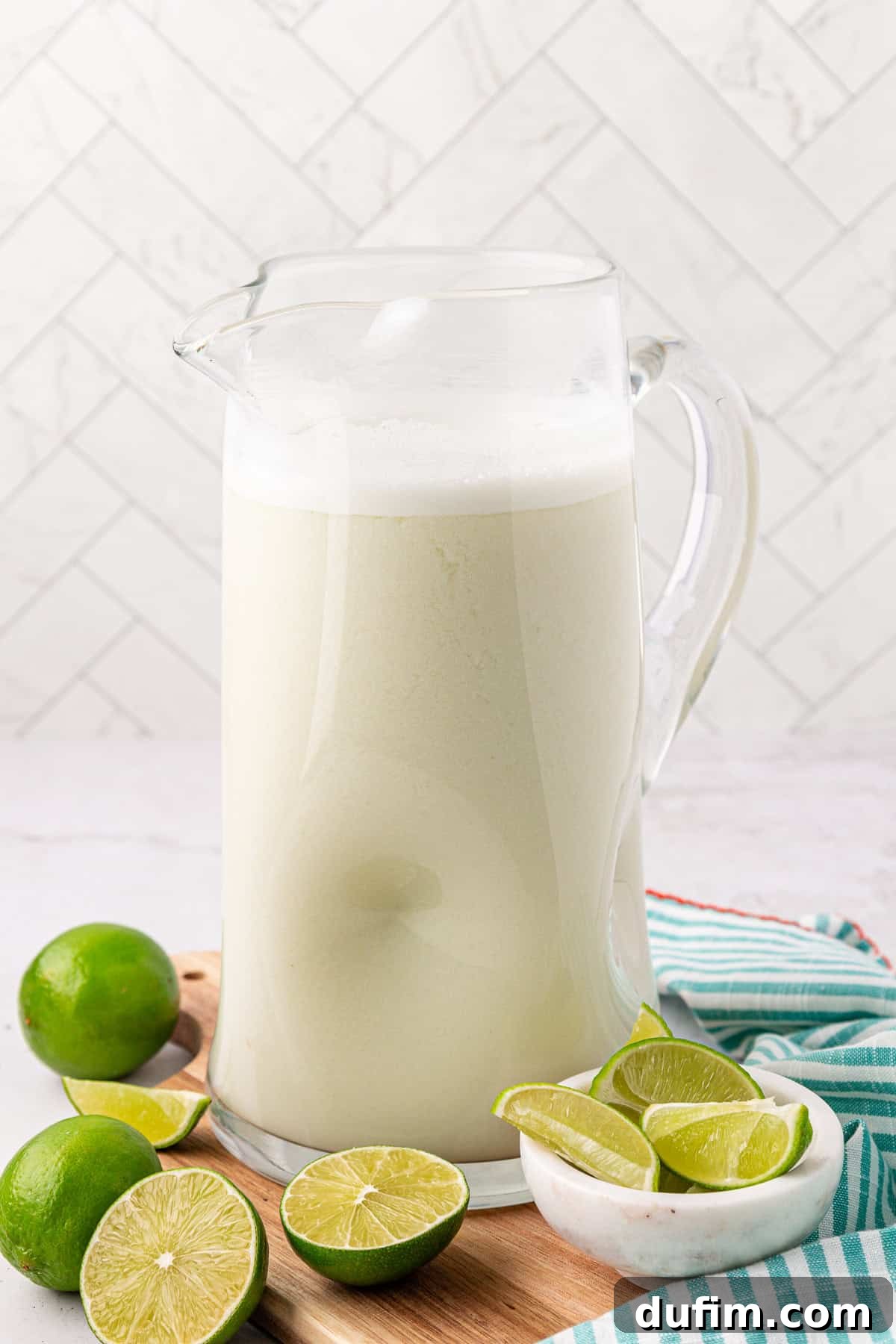 Crafting Creamy Brazilian Lemonade 10 A glass pitcher filled with a frothy, creamy white beverage sits on a wooden board, surrounded by whole and halved limes. A striped cloth is nearby, and the background features white tiled walls.