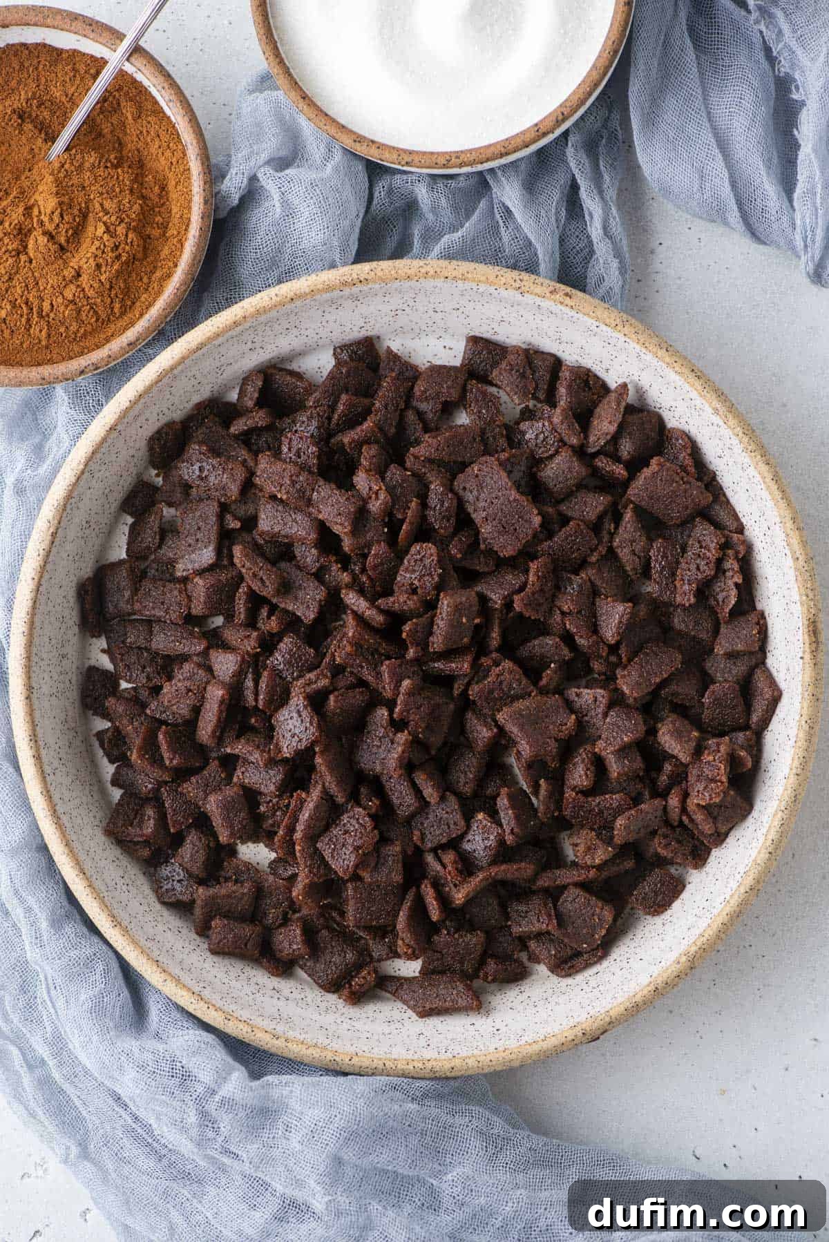 A bowl filled with rich, brown homemade cinnamon chips sits on a light gray surface, with small bowls of cinnamon powder and granulated sugar elegantly arranged nearby. A soft blue cloth provides a comforting backdrop.