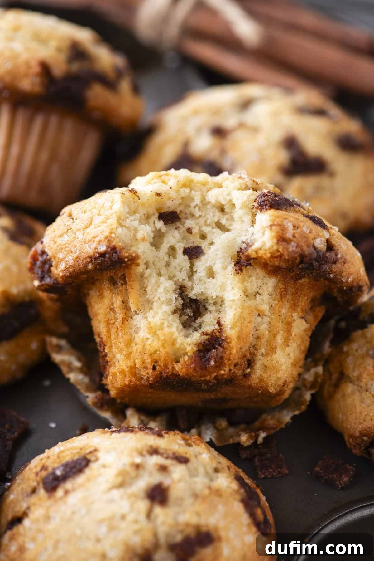A close-up of beautifully baked cinnamon chip muffins, with one muffin cut in half to showcase its moist, fluffy texture and generous chunks of melted cinnamon chips inside. Other whole muffins and cinnamon sticks are artfully arranged in the soft-focus background.