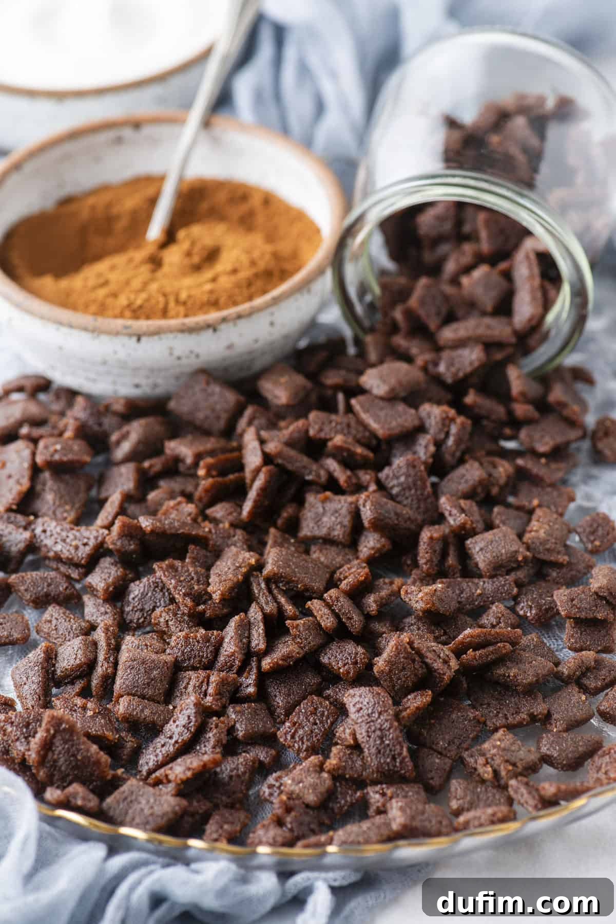 A baking tray neatly covered with freshly made cinnamon chips, positioned beside an overturned jar and a bowl of cinnamon powder containing a spoon. A light blue cloth is draped in the background, adding a touch of color.