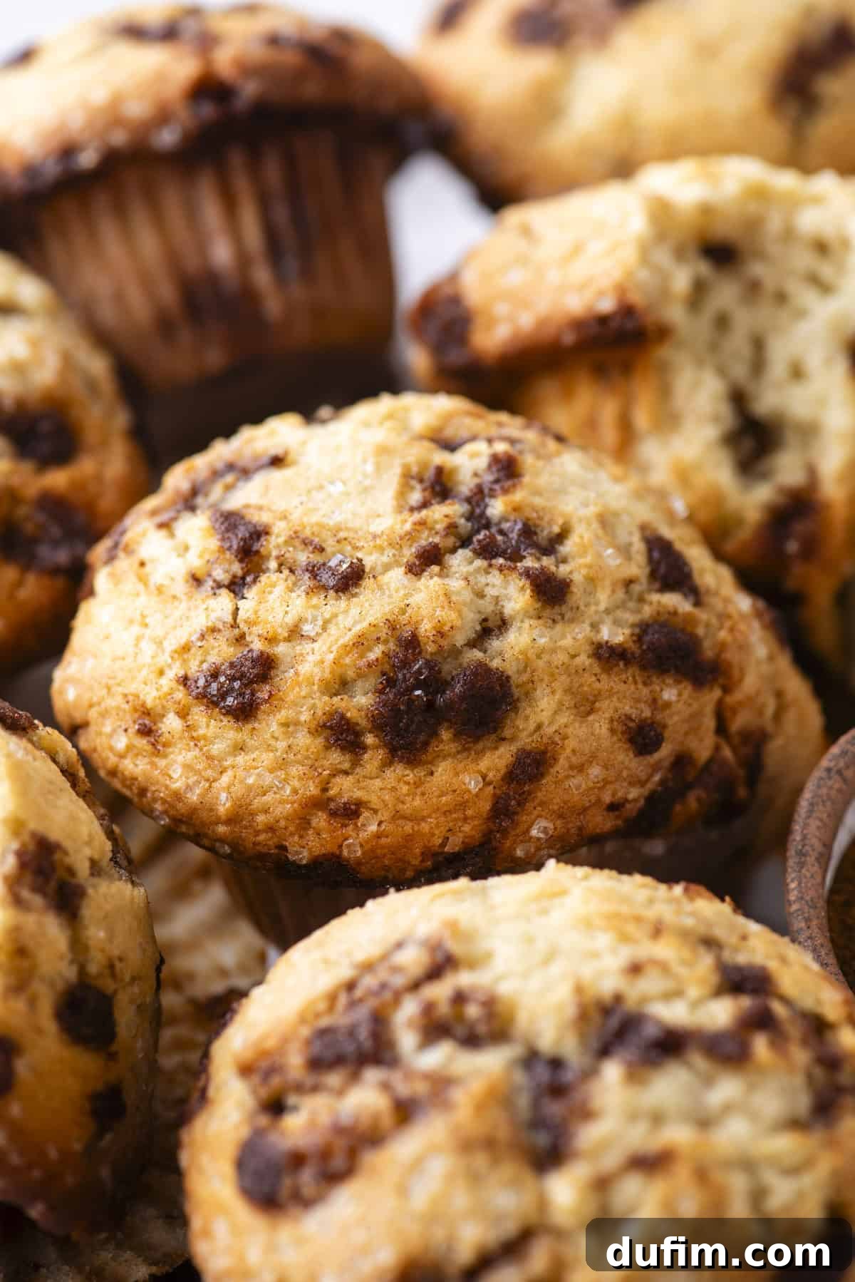 Close-up of golden, perfectly baked cinnamon chip muffins with slightly crisp tops. One muffin is partially broken open, revealing its soft, moist interior dotted with melted cinnamon pieces.