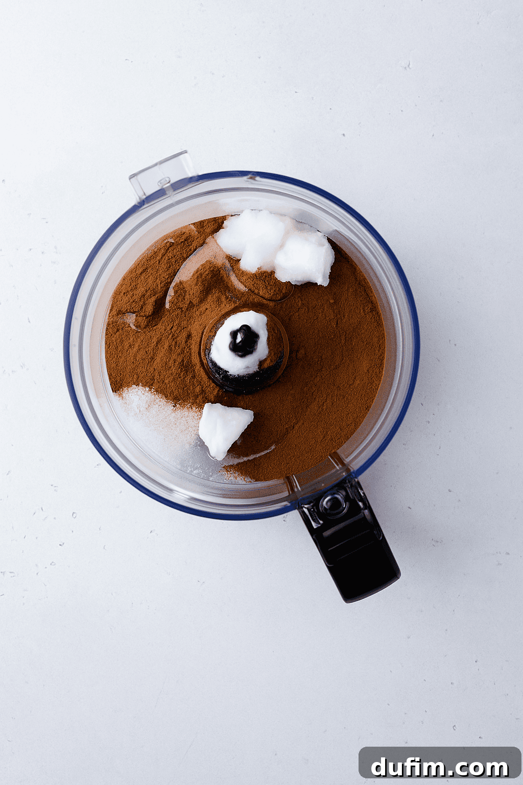 A food processor bowl containing ground cinnamon, granulated sugar, and several dollops of solid coconut oil on a light gray surface, illustrating the initial stage of ingredient mixing.