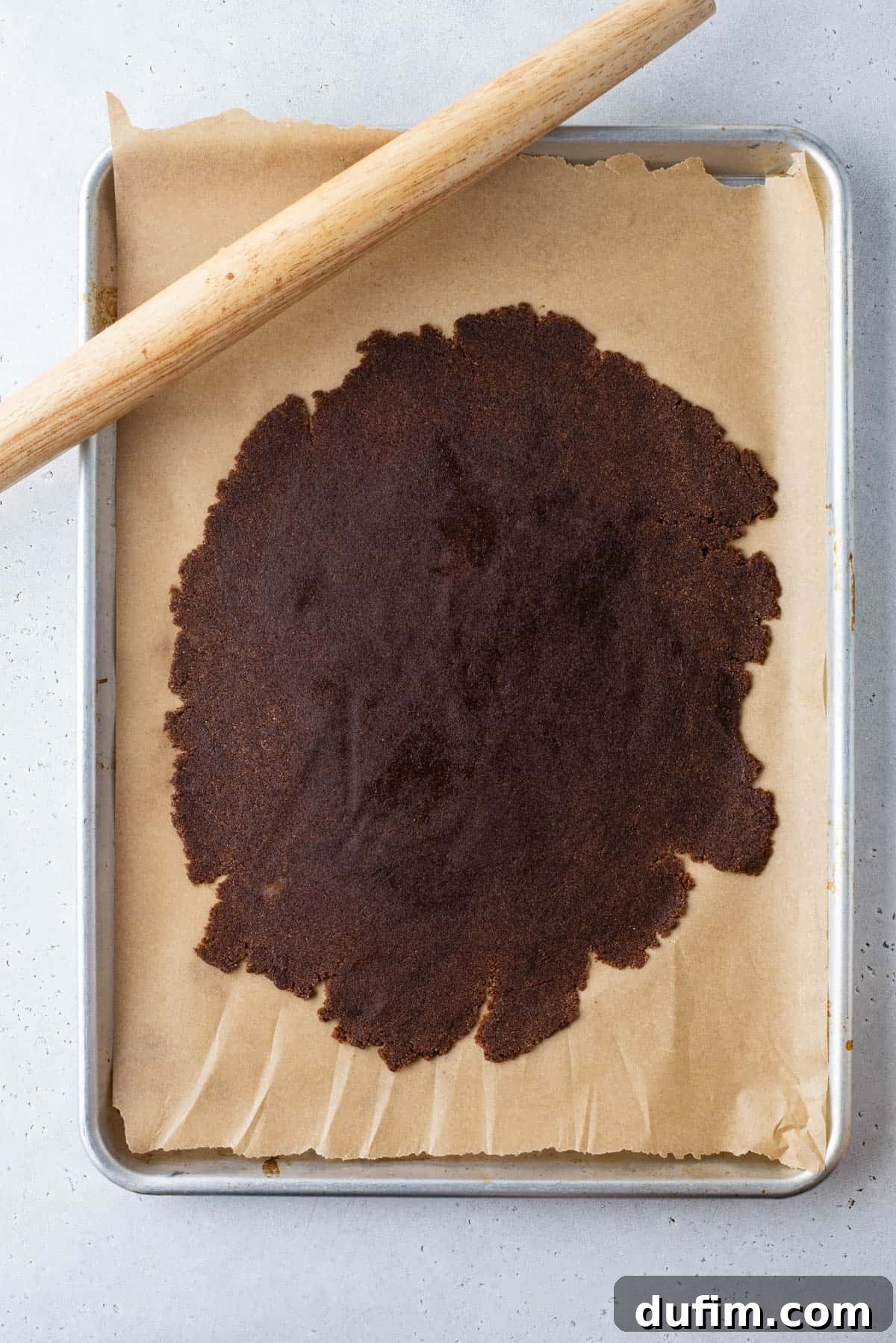 A baking tray lined with parchment paper holds a large, thin sheet of dark cinnamon dough, partially rolled out. A wooden rolling pin rests diagonally along the top edge of the tray, indicating the rolling process.