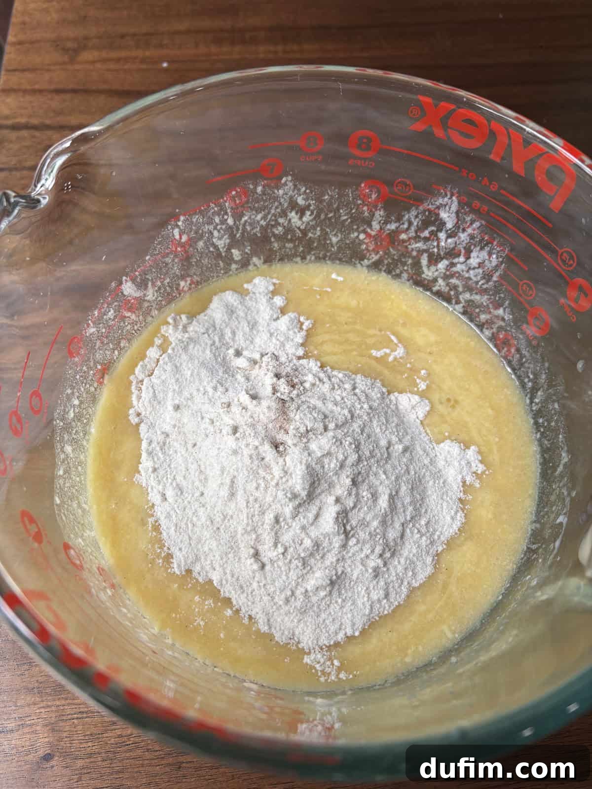 Spiced Cinnamon Chip Muffins 11 A glass Pyrex measuring cup containing a mixture of beaten eggs and sugar, with a mound of flour on top, sits on a wooden surface, ready to be mixed for baking.