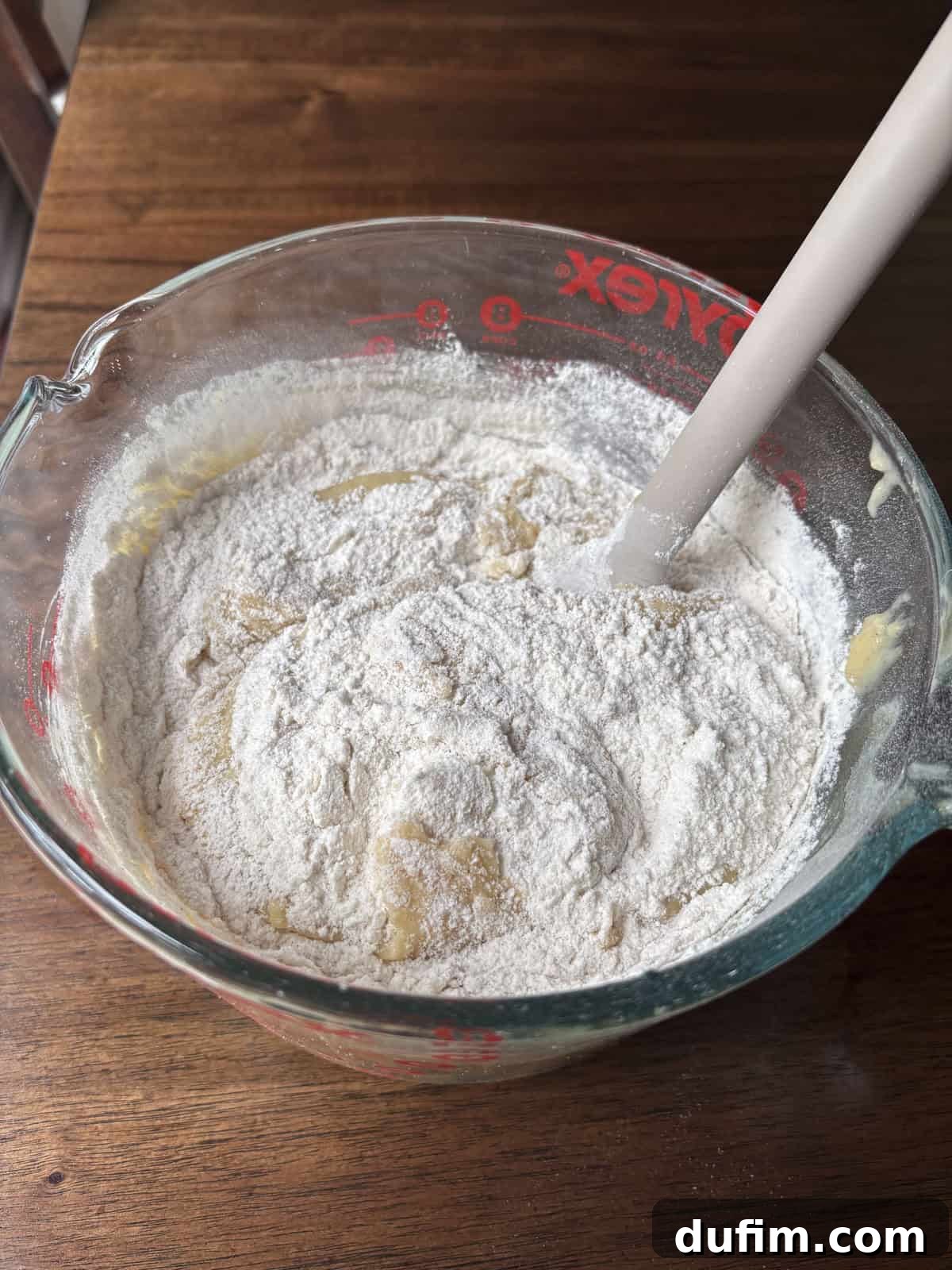 Spiced Cinnamon Chip Muffins 12 A glass mixing bowl filled with a mixture of flour and wet ingredients is being stirred with a white spatula on a wooden table. The flour is partially mixed into the batter.