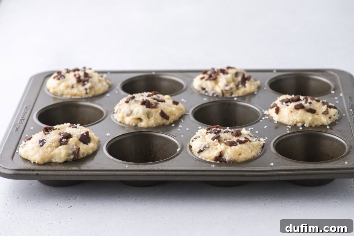Spiced Cinnamon Chip Muffins 14 A muffin tin with eight wells, four of which are filled with unbaked muffin batter topped with cinnamon chips and a sprinkle of salt, sits on a light gray surface.