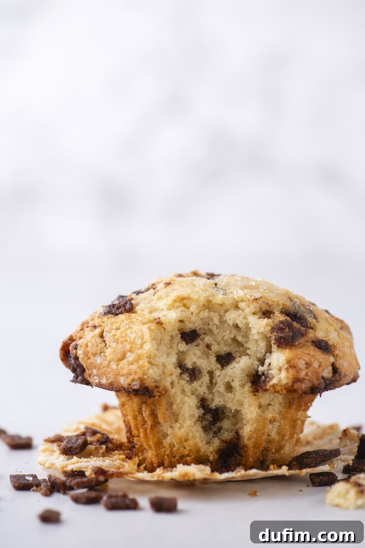 Spiced Cinnamon Chip Muffins 17 A cinnamon chip muffin with a bite taken out sits on a white surface, partially unwrapped from its paper liner. Crumbs and bits of cinnamon chips are scattered around.