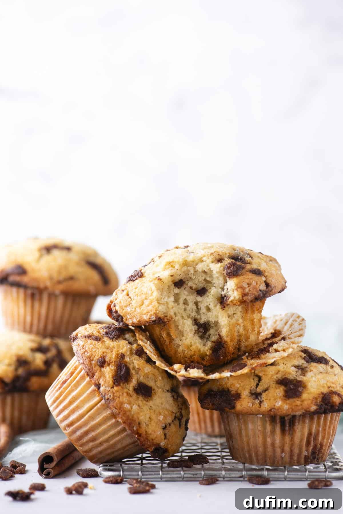 Spiced Cinnamon Chip Muffins 3 A stack of cinnamon chip muffins sits on a cooling rack, with one muffin cut open to show its soft, moist interior. The background is light and blurred, highlighting the muffins.