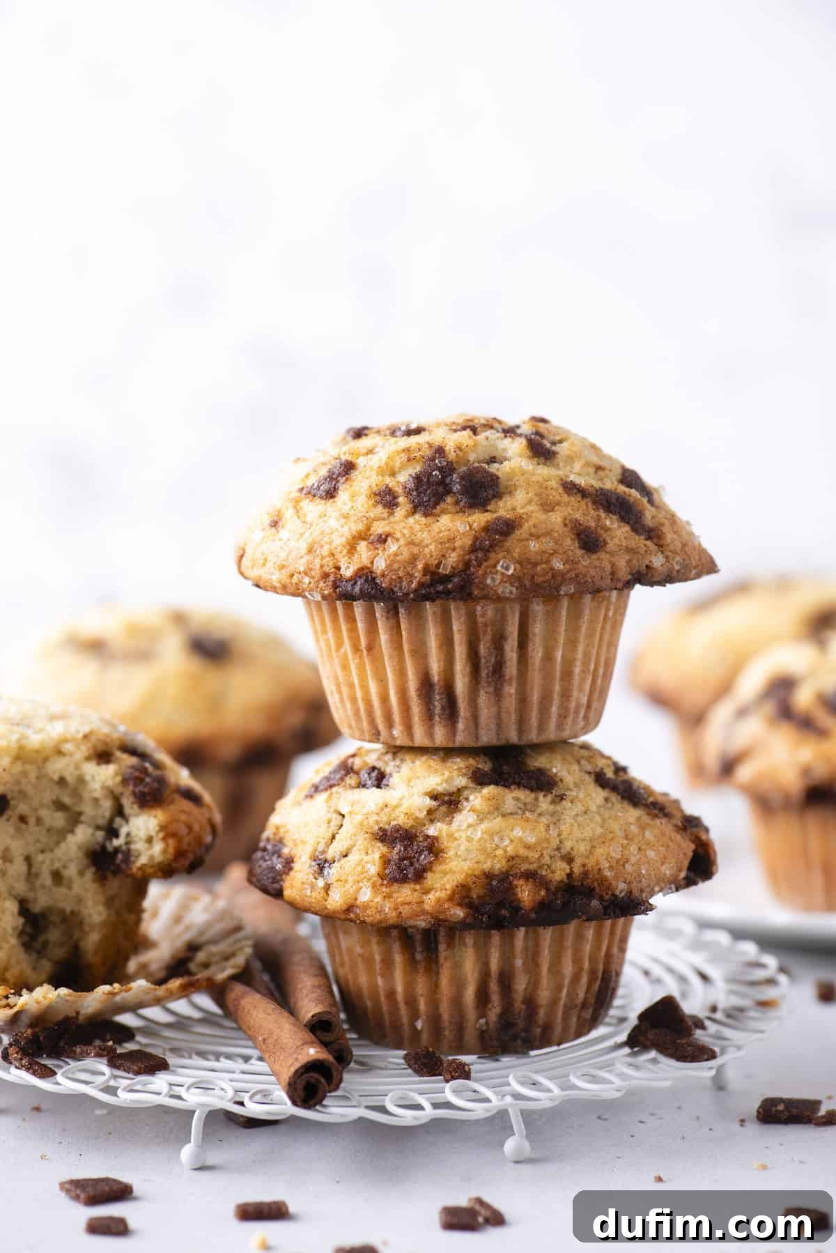 Spiced Cinnamon Chip Muffins 4 Two cinnamon chip muffins stacked on a white wire rack with additional muffins and loose cinnamon chunks in the background. A partially eaten muffin and cinnamon sticks are also visible.