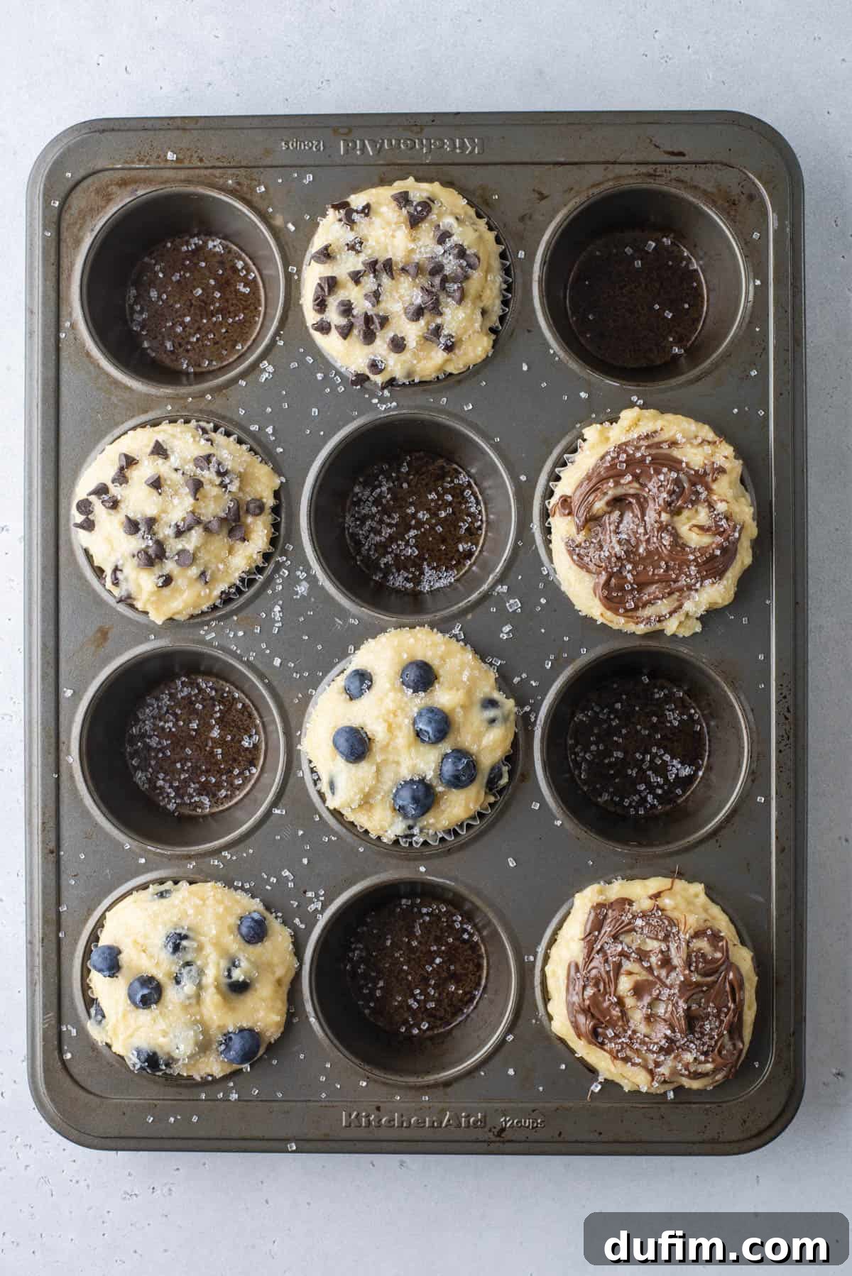 A muffin tin holds six unbaked muffins topped with chocolate chips, blueberries, or chocolate swirls, while three cupcake slots are empty, showing only the liners and sprinkled salt.