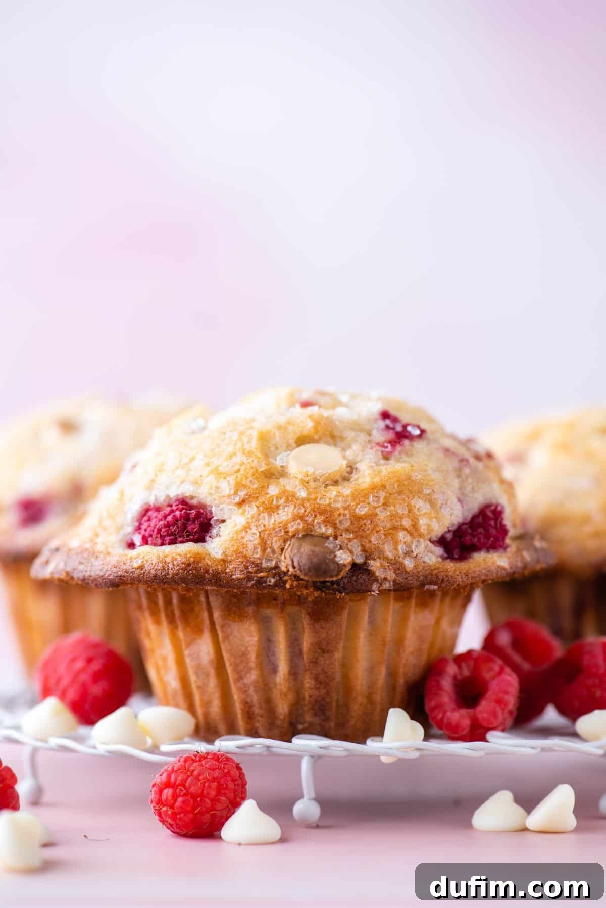 Close-up of a raspberry muffin topped with sugar crystals, surrounded by fresh raspberries and white chocolate chips on a white plate, with a soft pink background.