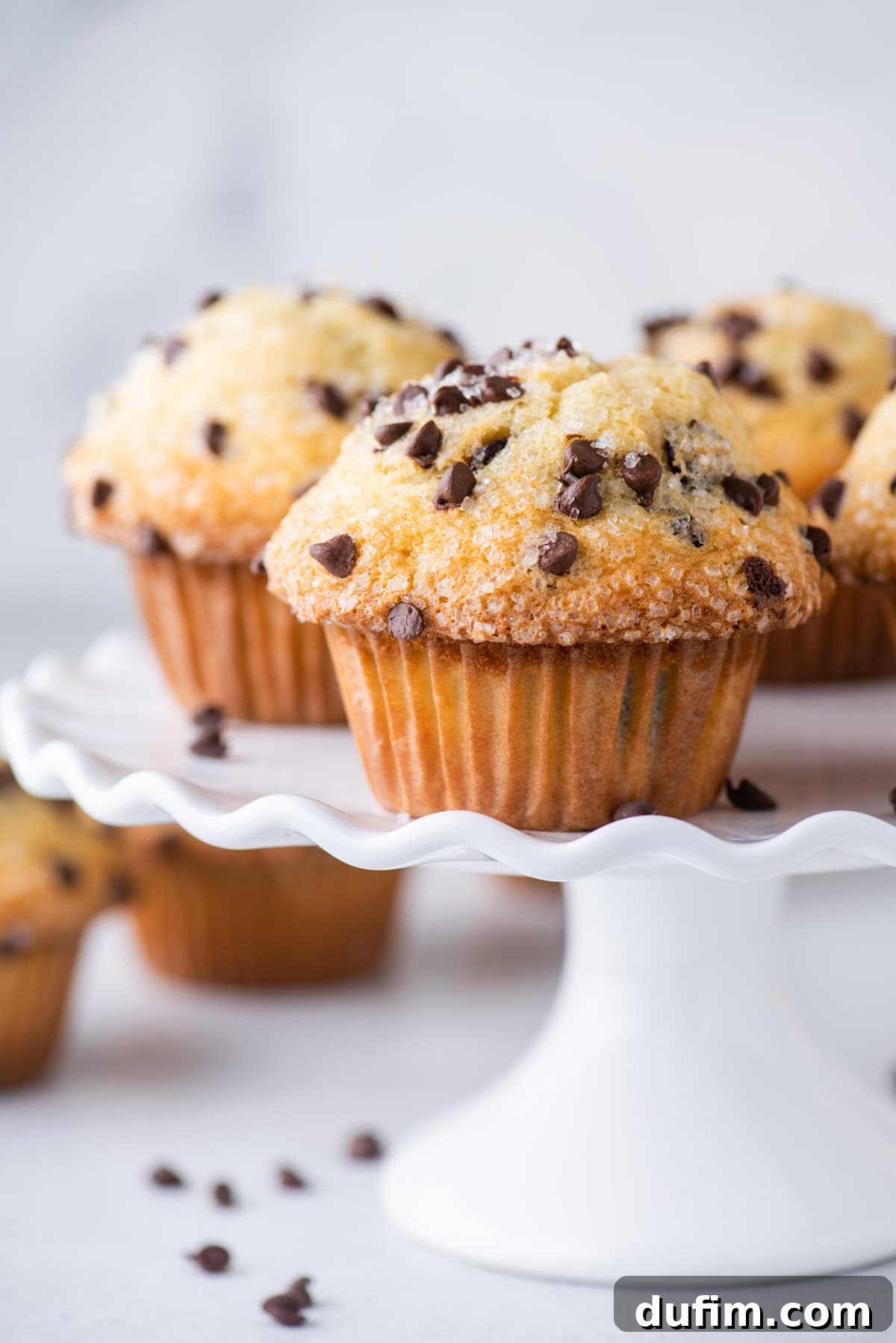 Chocolate chip muffins on a white, ruffled cake stand, with more muffins and scattered chocolate chips in the background on a light surface.