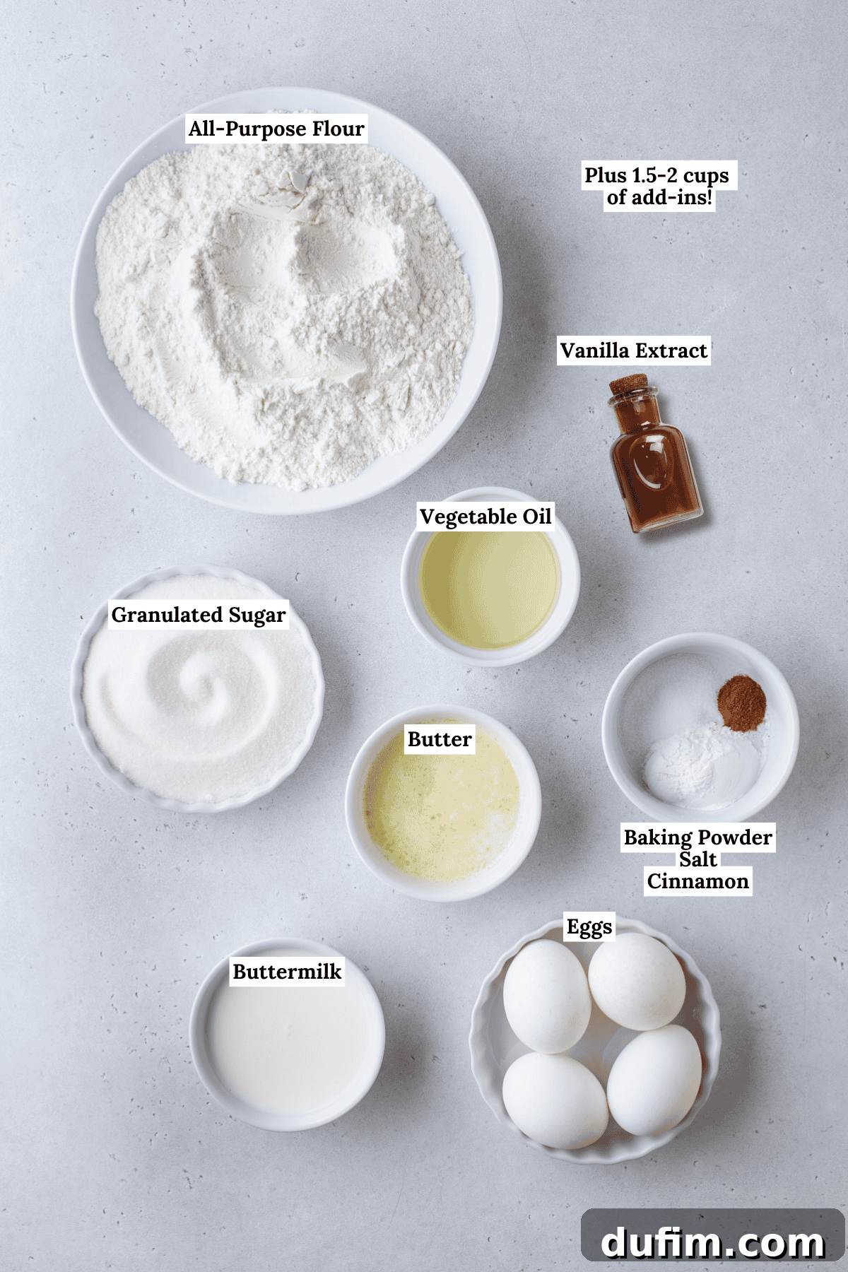 Top-down view of labeled baking ingredients in bowls: all-purpose flour, granulated sugar, buttermilk, eggs, melted butter, vegetable oil, vanilla extract, and a small bowl with baking powder, salt, and cinnamon.