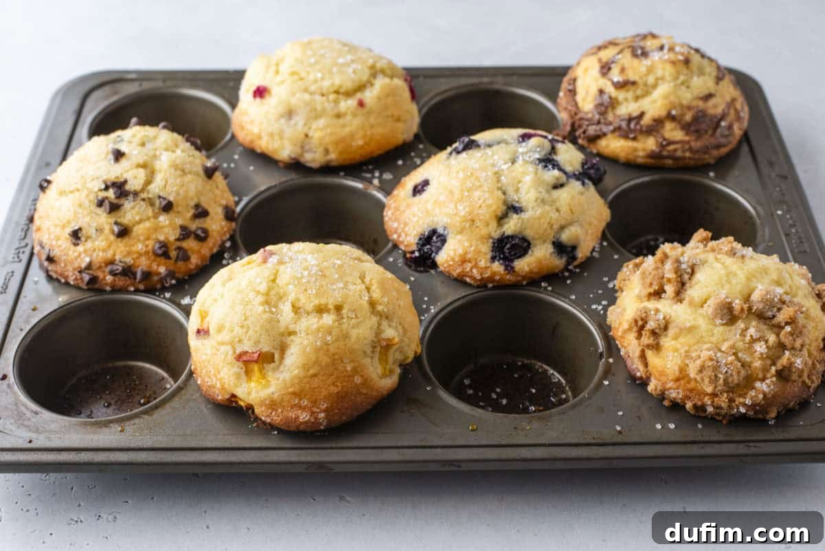 A metal muffin tray holding six assorted muffins, including ones with chocolate chips, blueberries, and crumble toppings, on a light gray surface.