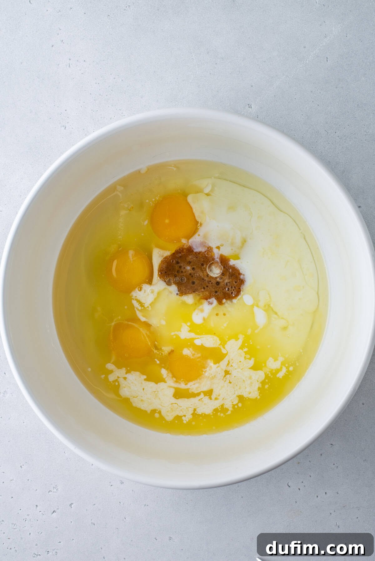 A white mixing bowl on a gray surface containing oil, vinegar, spices, and seasonings before being mixed together.