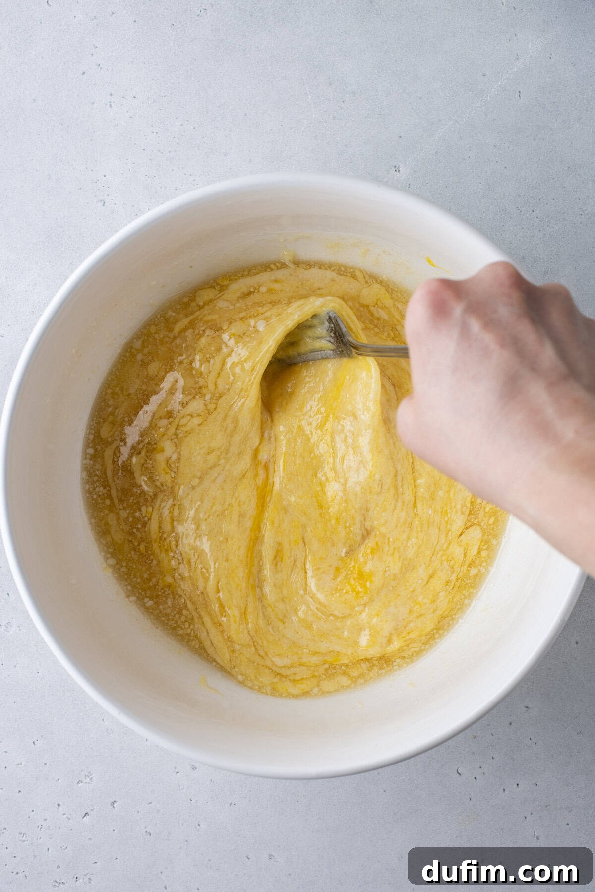 A hand uses a fork to mix yellow batter with specks of black seasoning in a white bowl on a light gray surface.