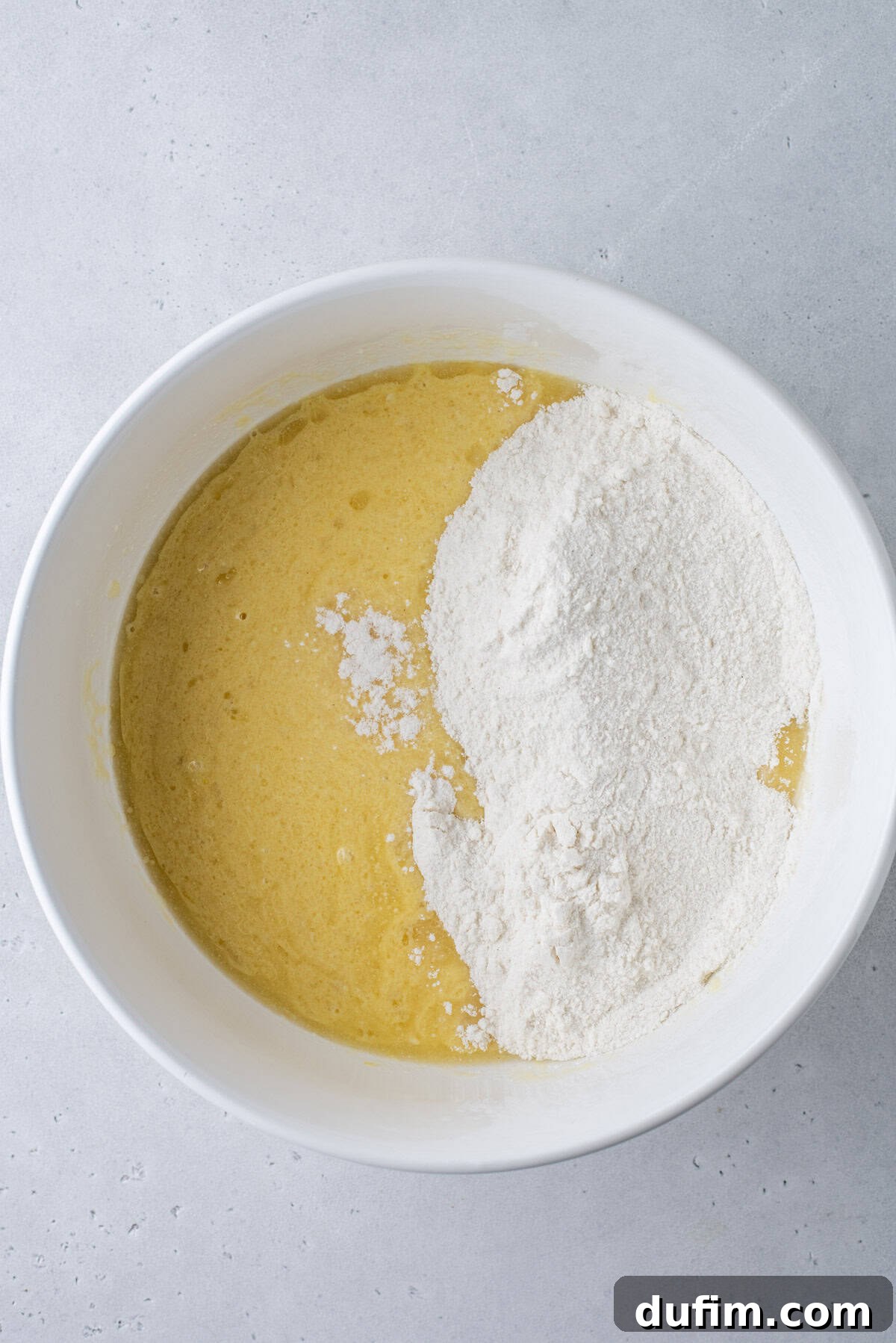 A white bowl containing a yellow liquid batter and a pile of white flour, partially mixed together, sits on a light gray surface.