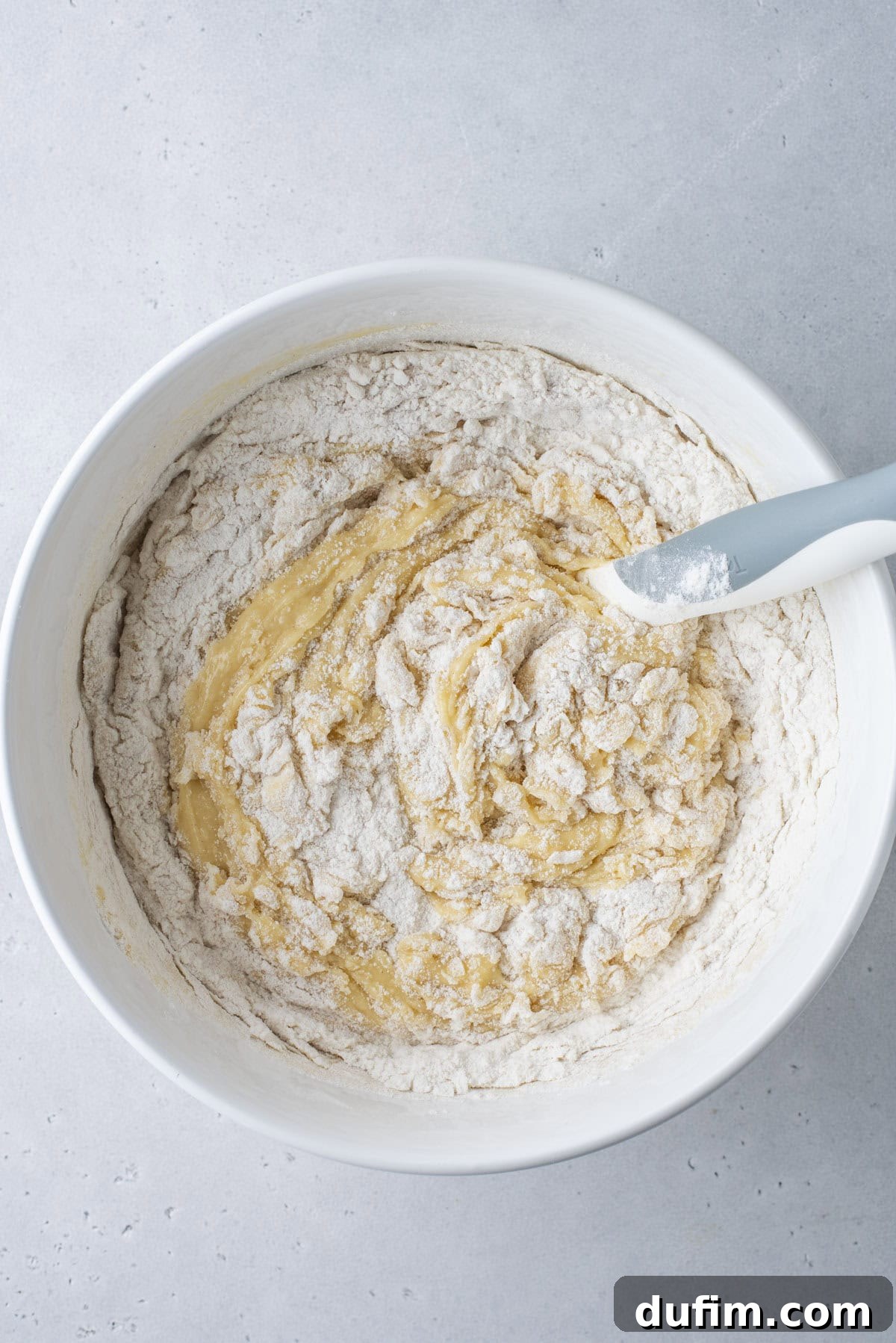 A white bowl containing partially mixed dough with visible dry flour and wet ingredients, and a gray spatula resting on the side of the bowl, on a light gray surface.