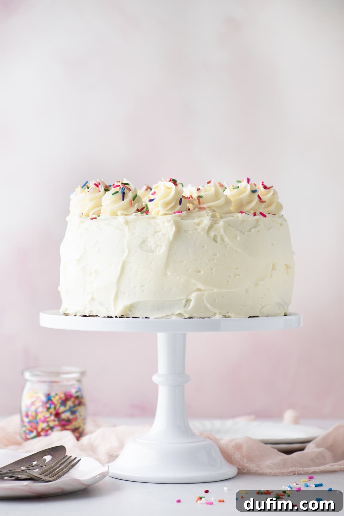 A beautifully round white frosted cake, elegantly topped with delicate swirls of frosting and a cascade of colorful sprinkles, rests on a pristine white cake stand. Nearby, a charming jar of sprinkles and a sleek cake server complete the inviting scene, all set against a soft, dreamy pink background.