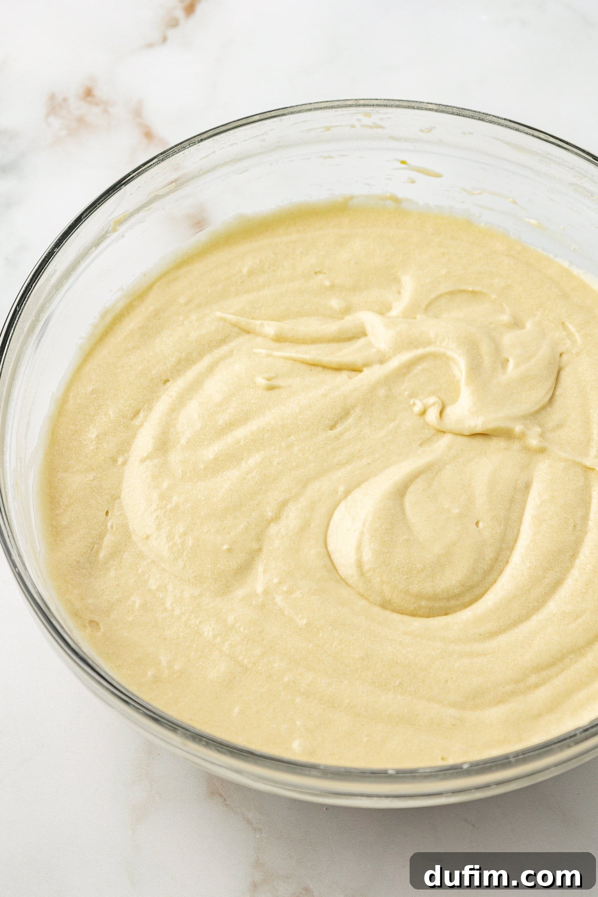 A glass bowl on a white marble surface contains a smooth, creamy, light yellow cake batter with subtle swirls on its surface.
