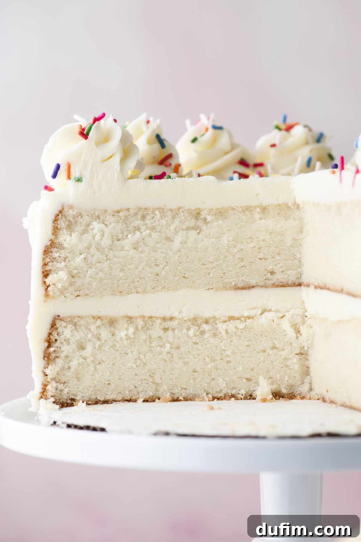 A close-up view of a beautifully frosted white layer cake, featuring two thick, moist layers, artfully topped with luscious swirls of frosting and a vibrant scattering of colorful rainbow sprinkles. It sits on a classic white cake stand, with a large slice already removed, revealing the soft, fluffy texture of the cake’s interior.