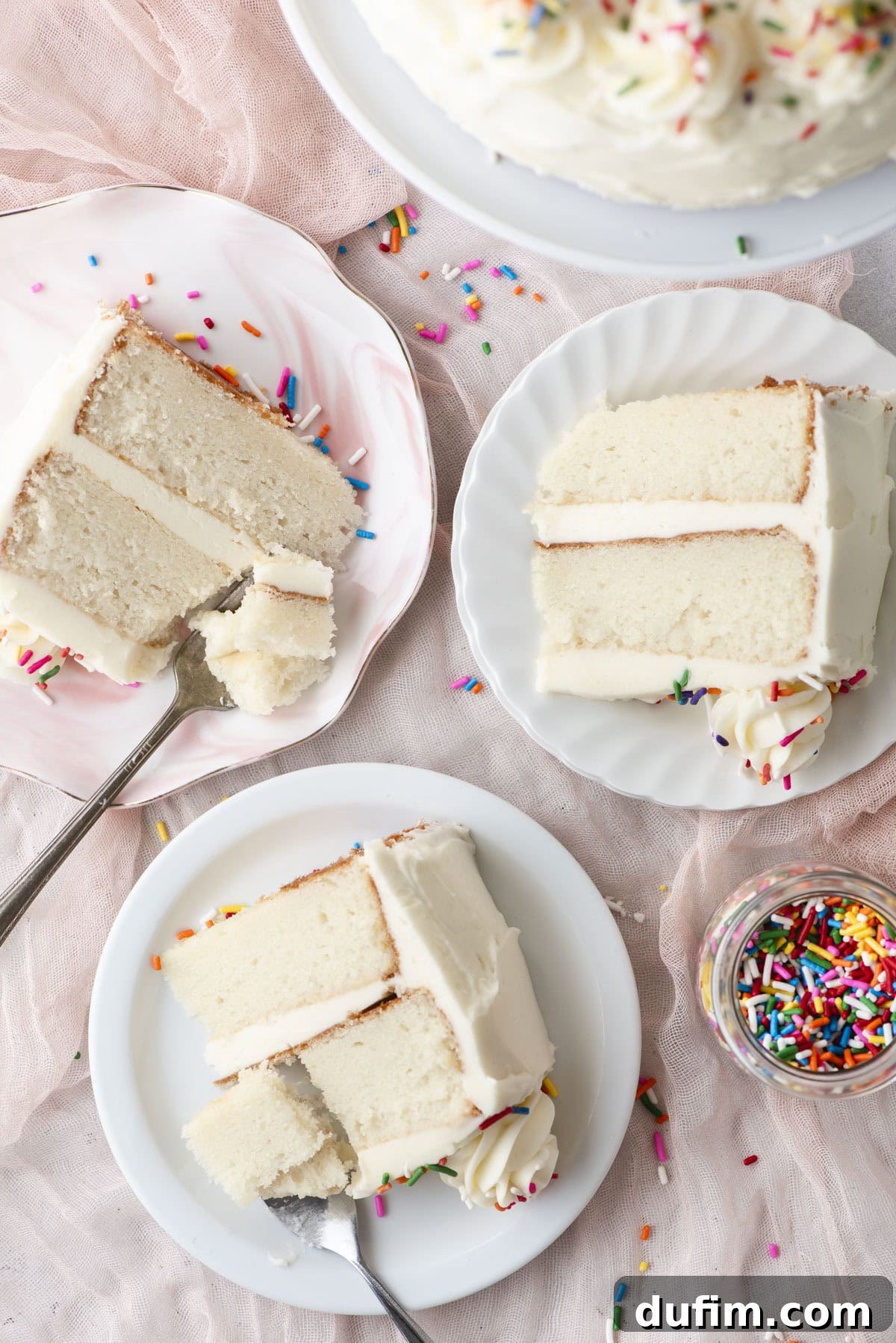 Three individual plates, each presenting a perfectly portioned slice of white cake generously topped with creamy white frosting and a joyful scattering of colorful sprinkles. A small, decorative jar of sprinkles and a soft pink cloth are artfully arranged nearby, completing a delightful and festive dessert tableau.