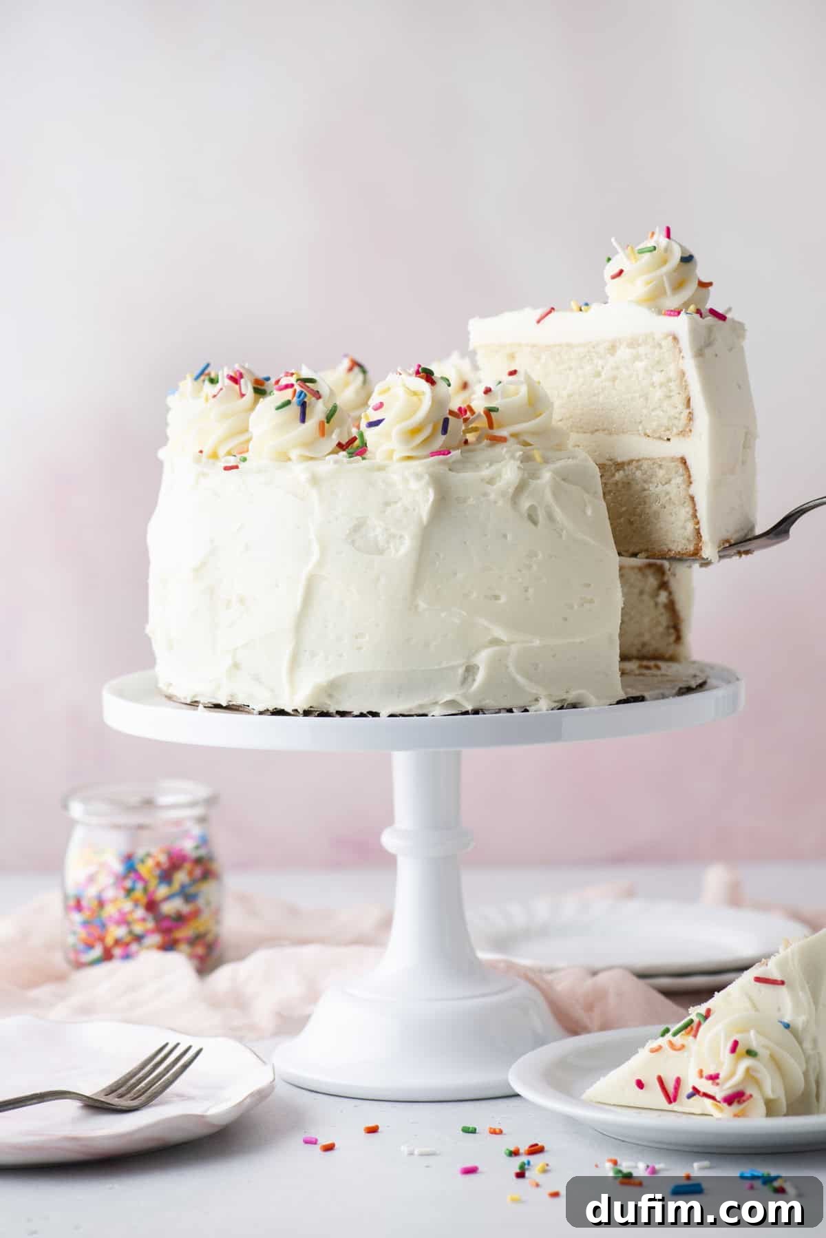 A white frosted layer cake, elegantly decorated with swirls of icing and a generous scattering of colorful sprinkles, is proudly displayed on a cake stand. A slice is being carefully lifted, revealing the soft, fluffy interior of the cake. Nearby, a whimsical jar of sprinkles, extra plates, and utensils are arranged, setting a festive and inviting scene.
