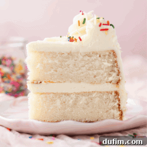 A close-up of a slice of white cake with two layers, filled and topped with white frosting and colorful sprinkles, on a light pink plate. A jar of sprinkles is blurred in the background.