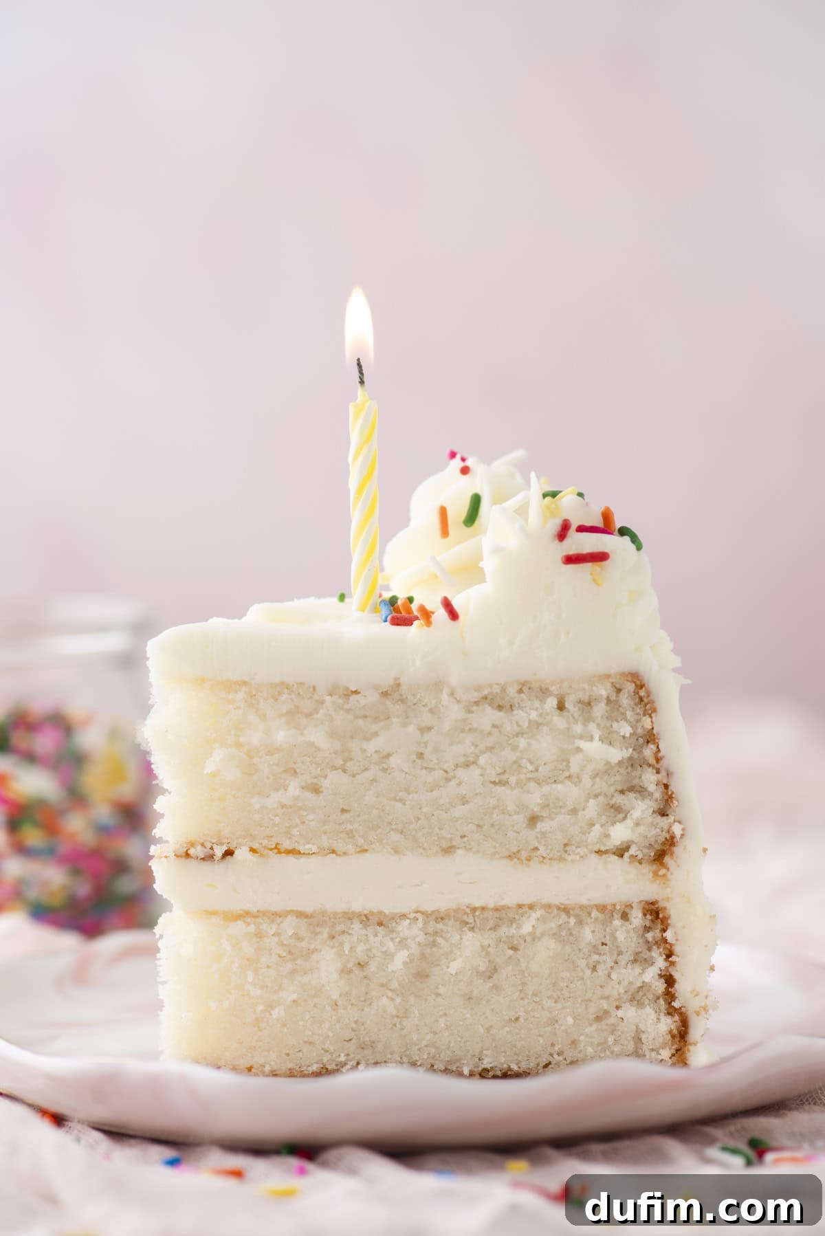 A delectable slice of white layer cake, generously adorned with creamy white frosting, rainbow sprinkles, and a charming lit yellow-striped candle, is presented on a plate. The background softly blurs, revealing a whimsical jar of sprinkles nearby, adding a festive touch to the dessert.