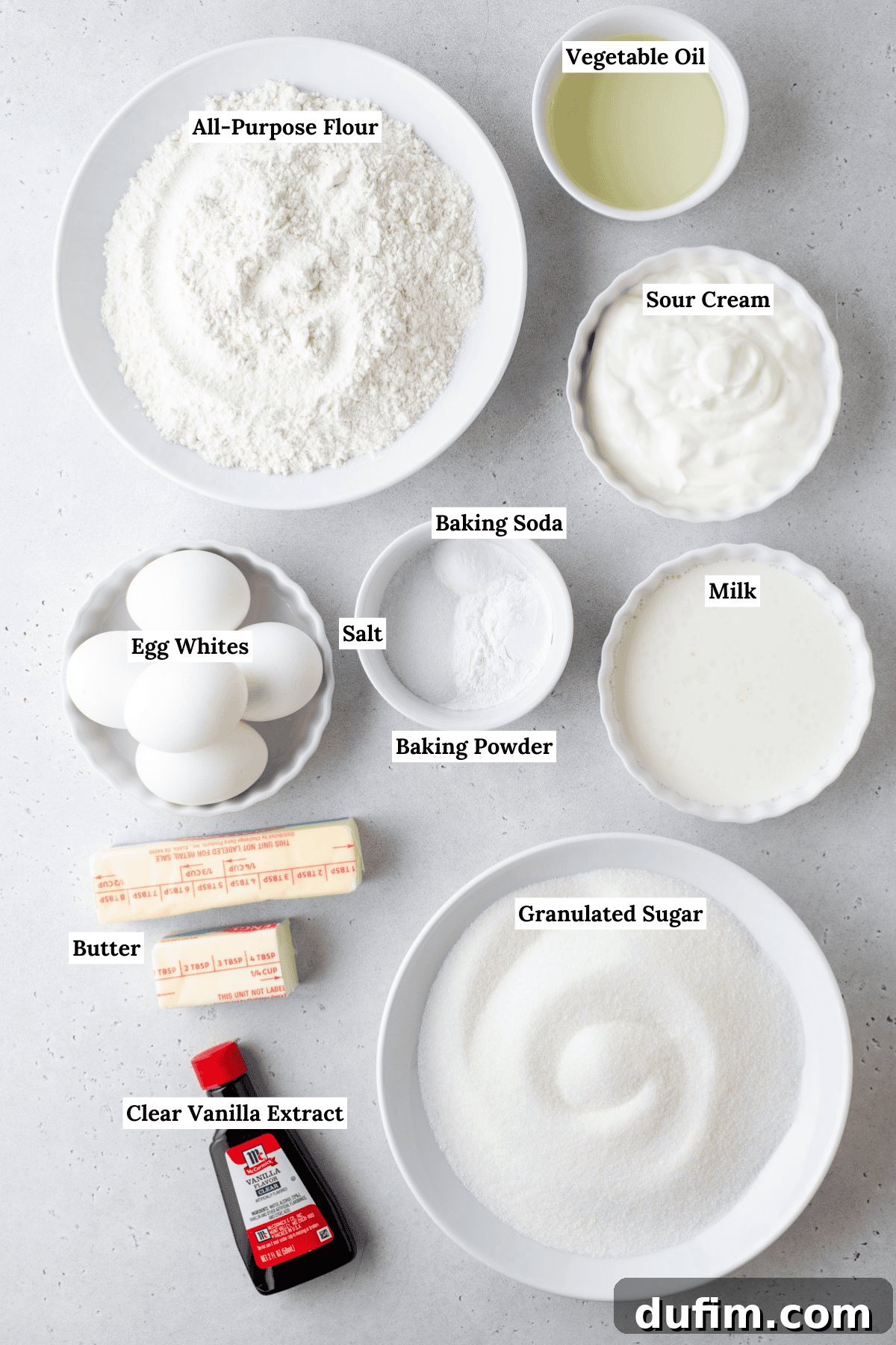A perfectly arranged overhead view showcases essential baking ingredients on a clean white surface. Labeled containers hold all-purpose flour, granulated sugar, sticks of butter, eggs, vanilla extract, vegetable oil, sour cream, milk, baking powder, baking soda, and salt, ready for creating a delicious cake.