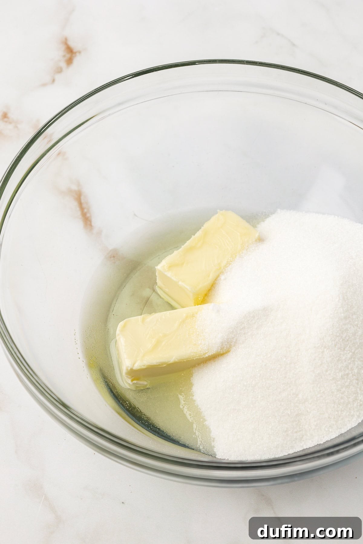 Two sticks of softened butter and a mound of sparkling granulated white sugar sit in a clear glass bowl on a pristine marble surface, ready for creaming.