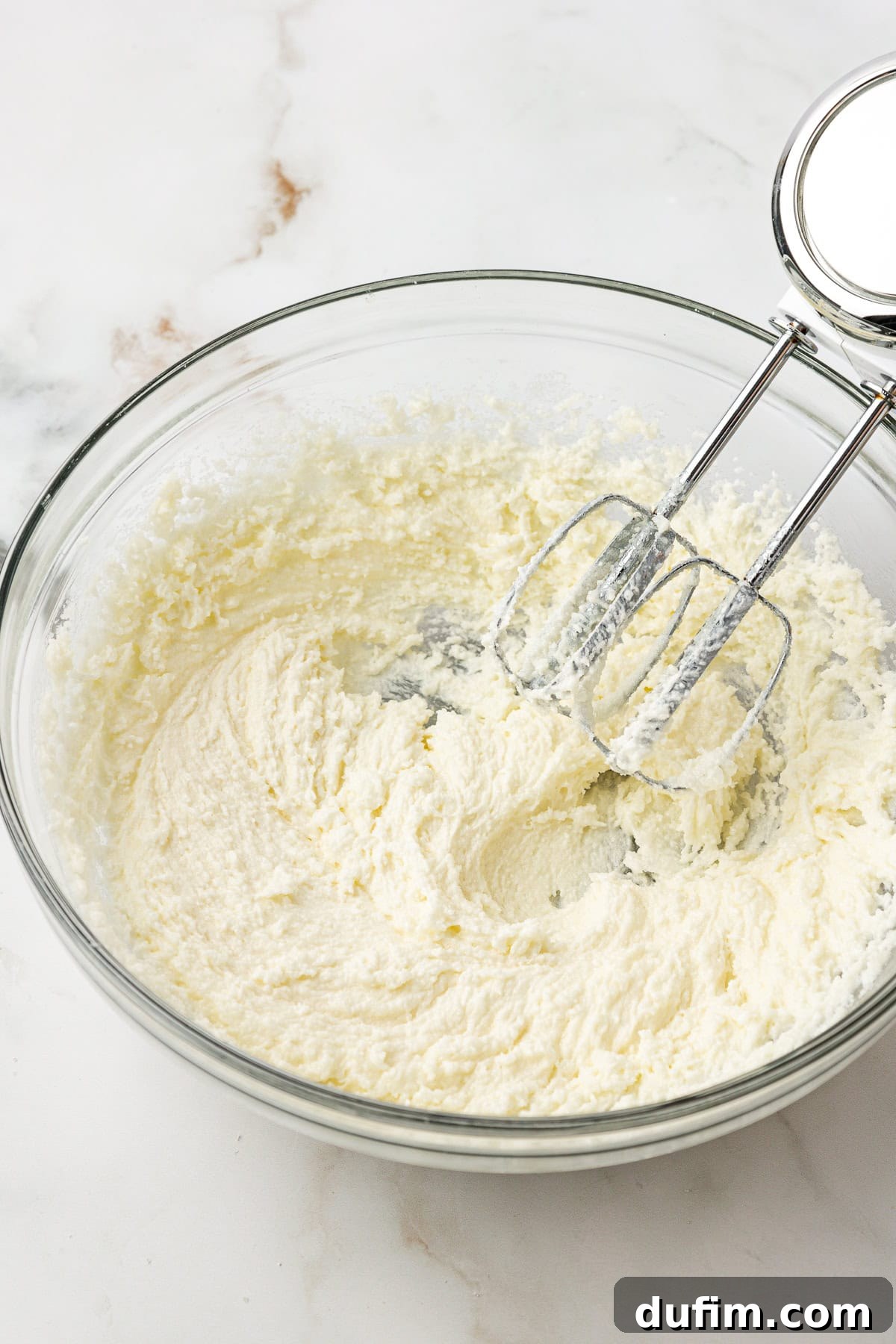An electric hand mixer with two beaters rests inside a glass bowl filled with a creamy, light yellow mixture of butter and sugar, positioned on a white marble surface.