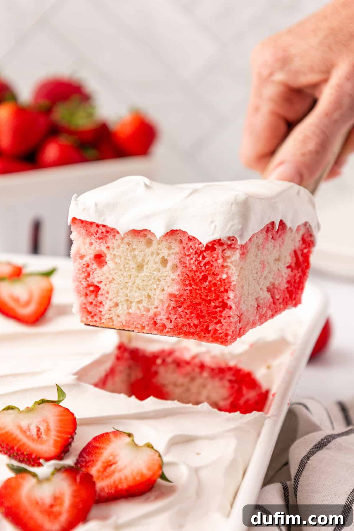 A hand lifts a slice of white and red poke cake topped with whipped cream from a tray. Fresh strawberries are on the tray and in a bowl in the background, highlighting the cake's vibrant colors and fresh garnishes.