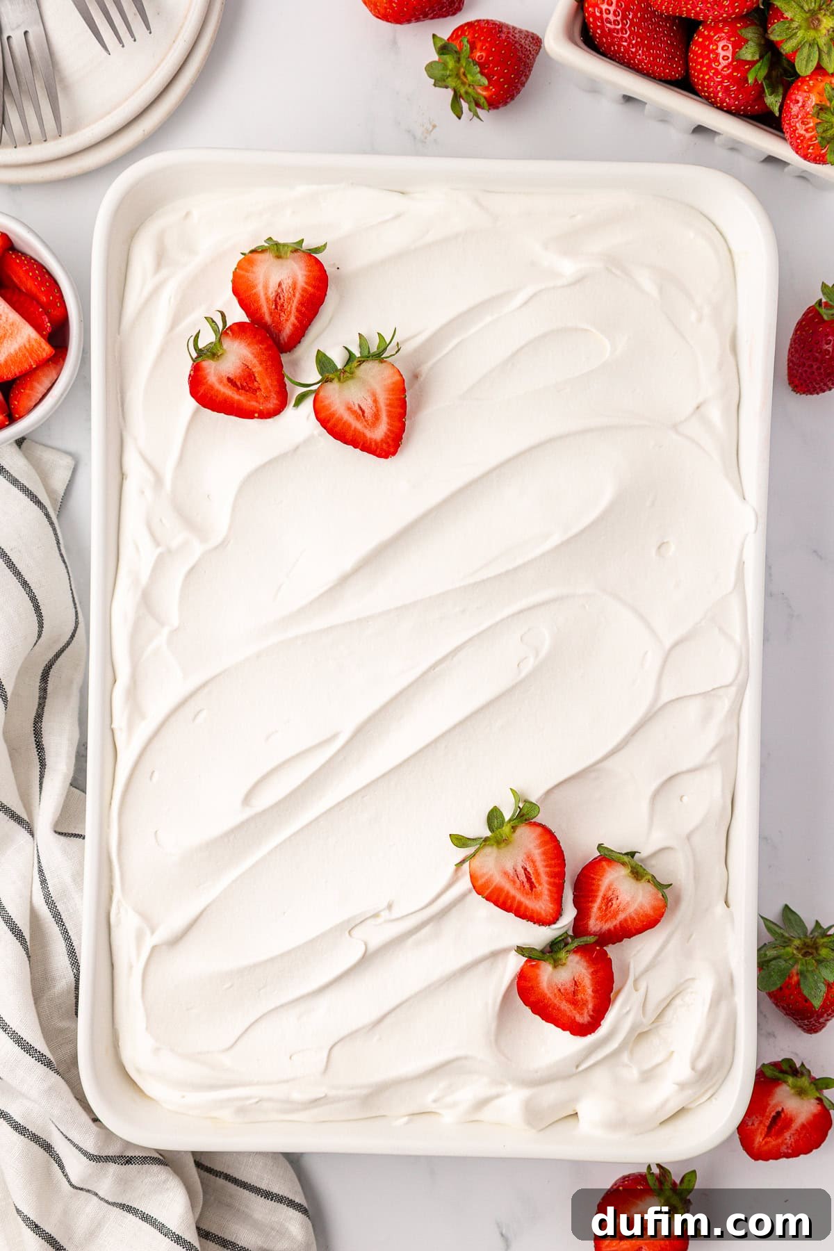 A rectangular dish filled with a dessert topped with smooth, white whipped cream, garnished with fresh, halved strawberries. Whole and sliced strawberries are scattered nearby on a white surface, ready for serving.