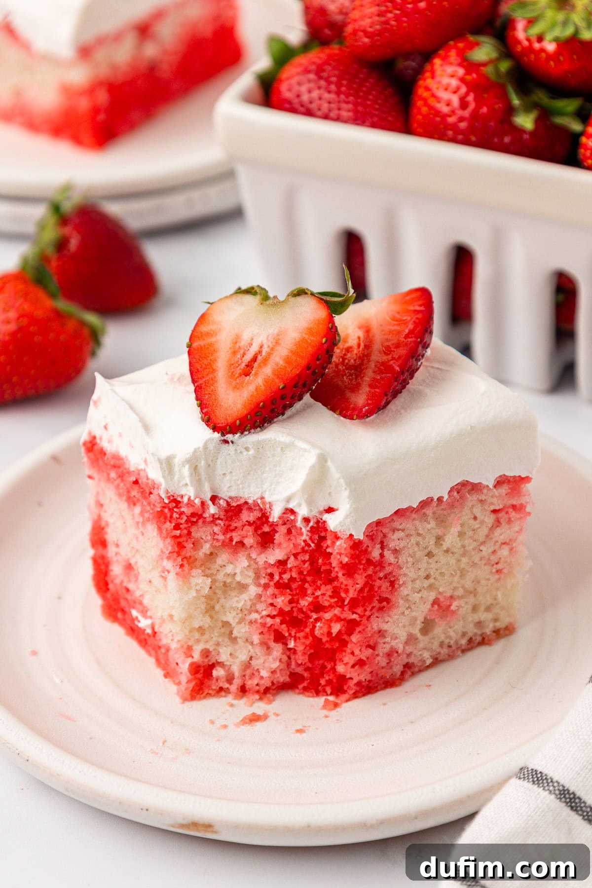 A slice of strawberry poke cake with white frosting and two strawberry halves on top, served on a plate with fresh strawberries and a container of strawberries in the background, showcasing its delicious layers.