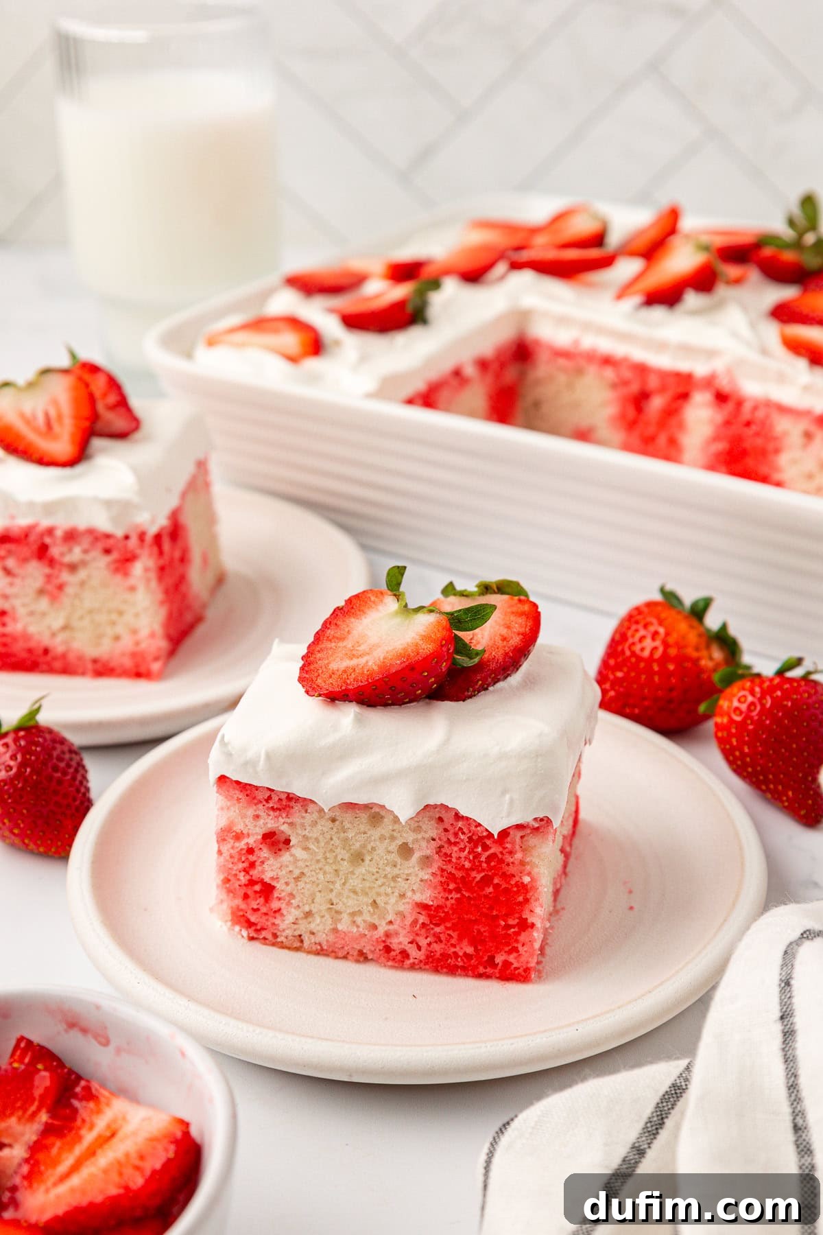 A slice of strawberry poke cake topped with whipped cream and fresh strawberry slices on a plate, with more cake and strawberries in the background, ready to be enjoyed.